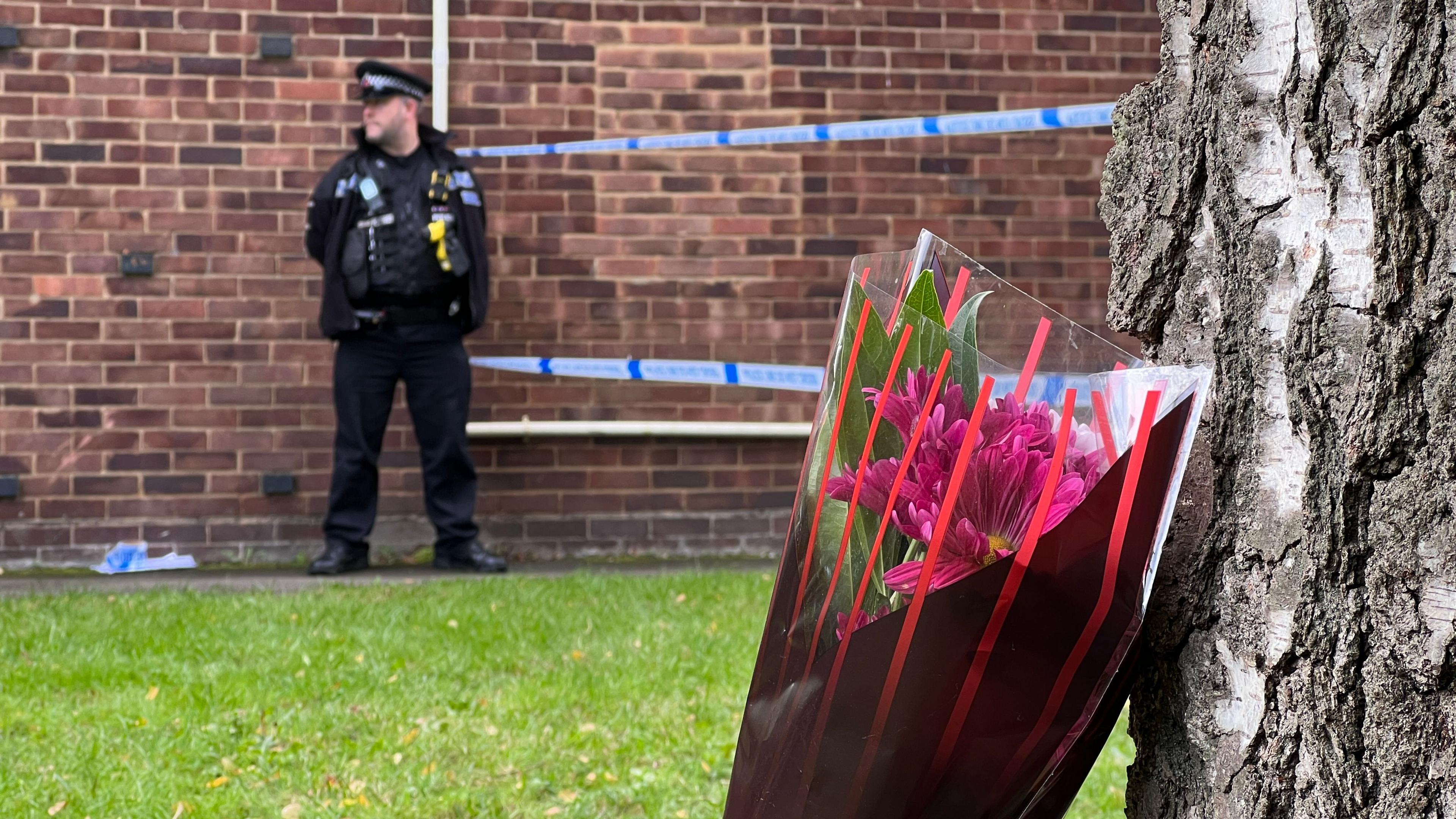 A bouquet of flowers left by a tree. In the photo there is also a police officer stood by a police cordon. 