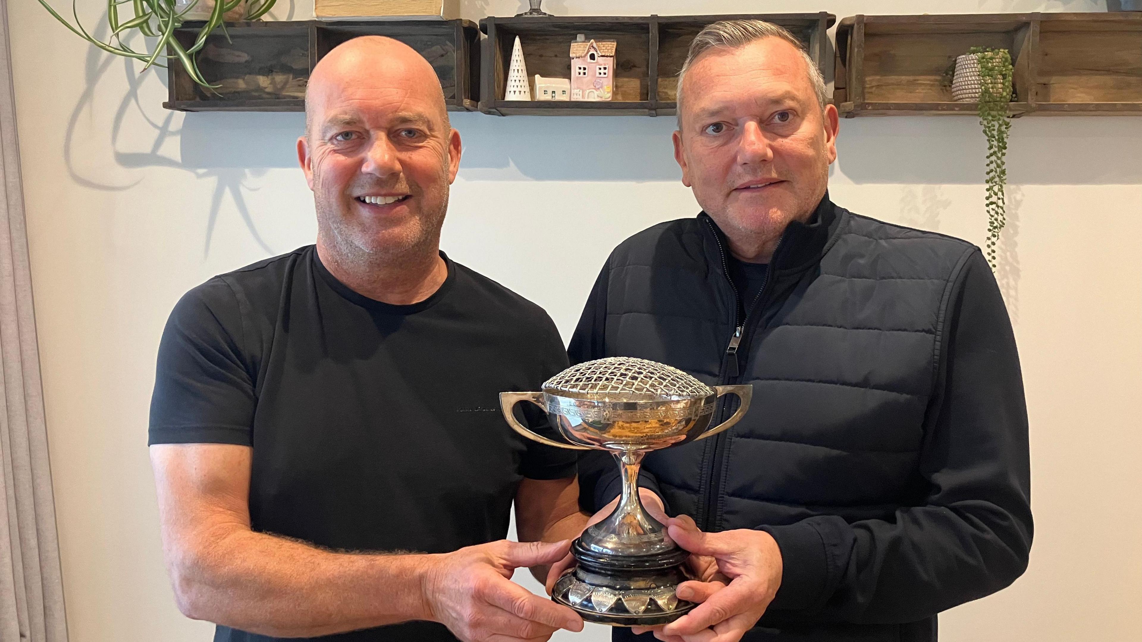 Two men hold a silver cup trophy. The man on the left has a bald head, and is wearing a black T shirt. The man on the right has short grey hair and is wearing a black jacket. They are standing against a white wall with brown shelves.