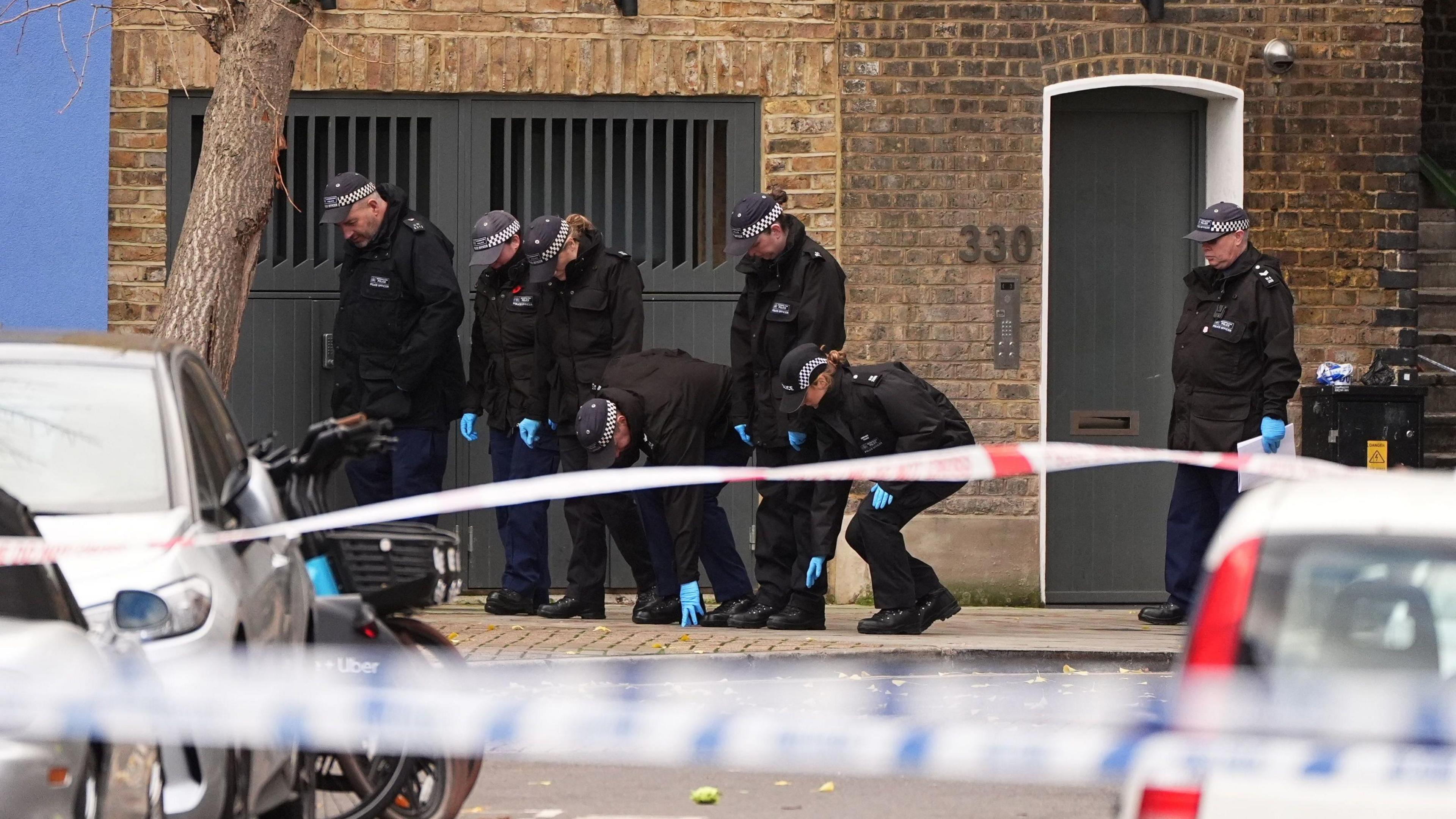 Officers in blue gloves, black uniforms and black caps with a checked strip proceed with a fingertip search in a line along the pavement, which is behind a cordon