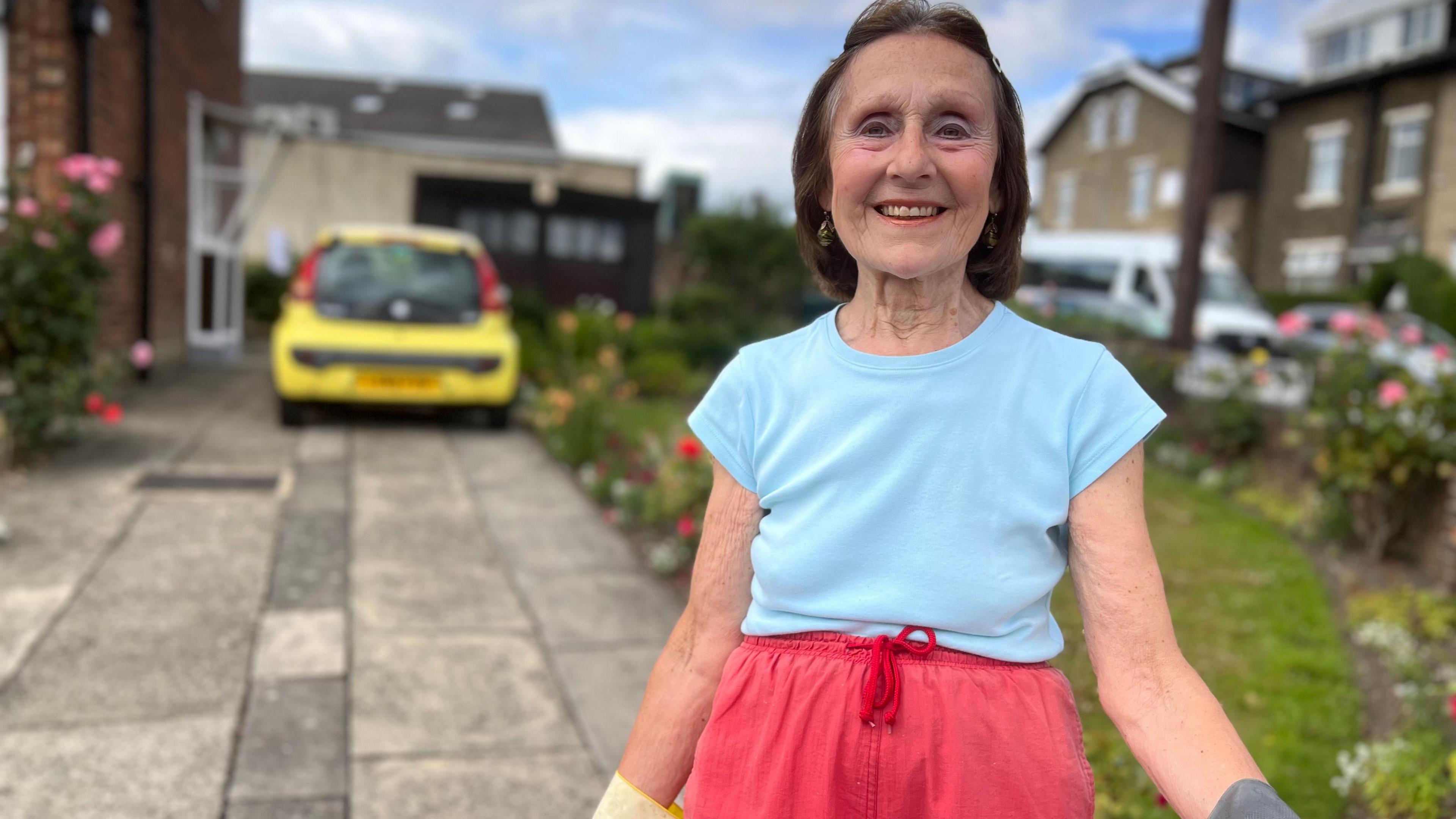 An older lady standing in her front garden and driveway. She wears gardening gloves. There is a car parked behind her and there are flowers and grass verges around her which look pretty and well kept.