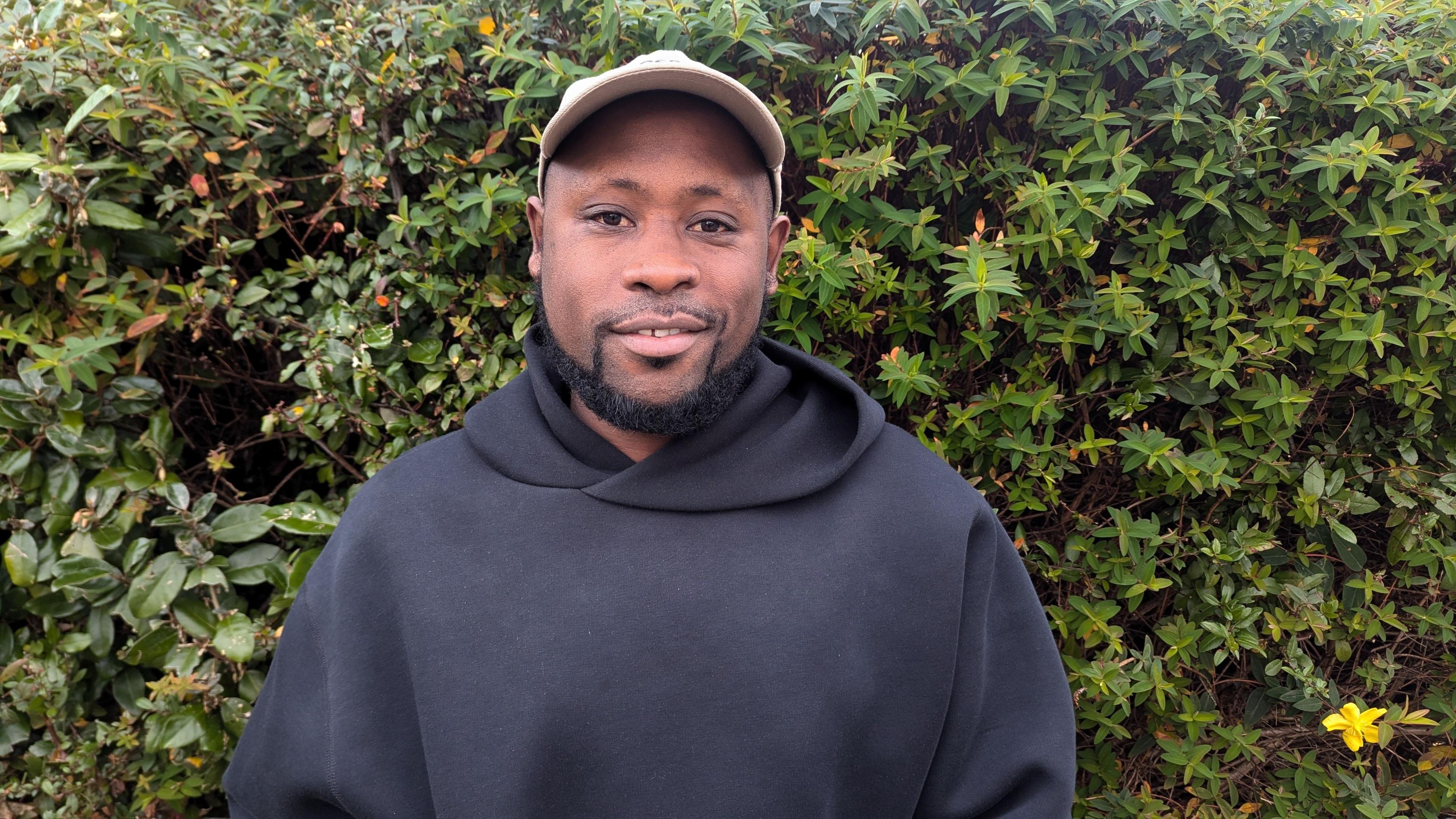 Rayan Wilson. He has a short black beard. He is wearing a black hoodie and a cream cap. He is pictured in front of a green hedge, looking directly at the camera and smiling.