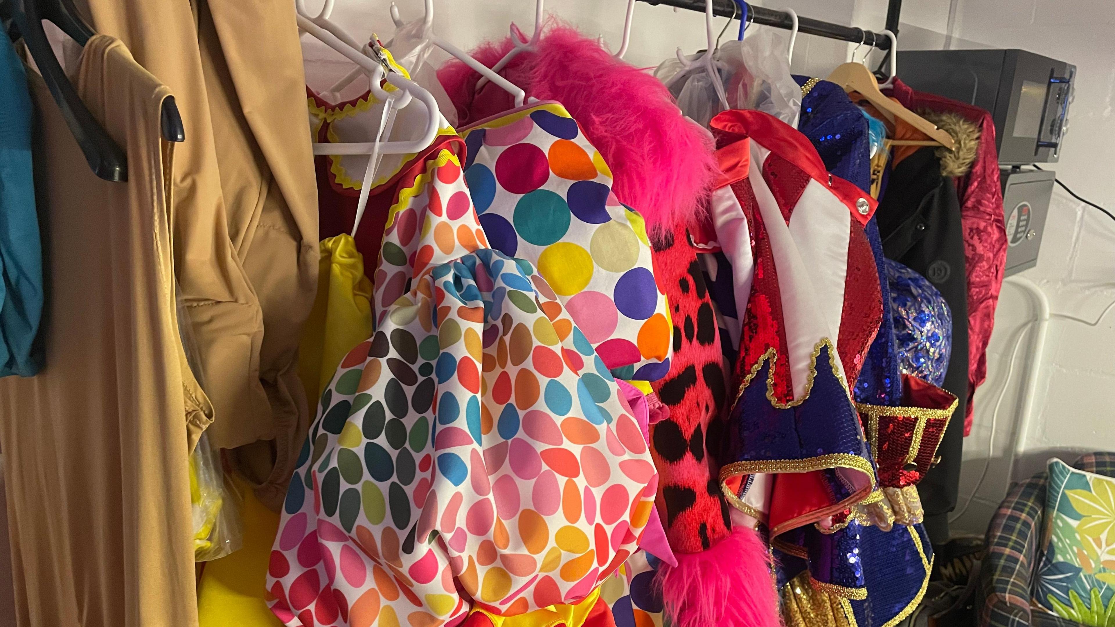 A row of colourful costumes on a rack in a dressing room, on hangers. They have fur and sequins on them, with a chair to the right. There is a lighting box to the right, higher up.