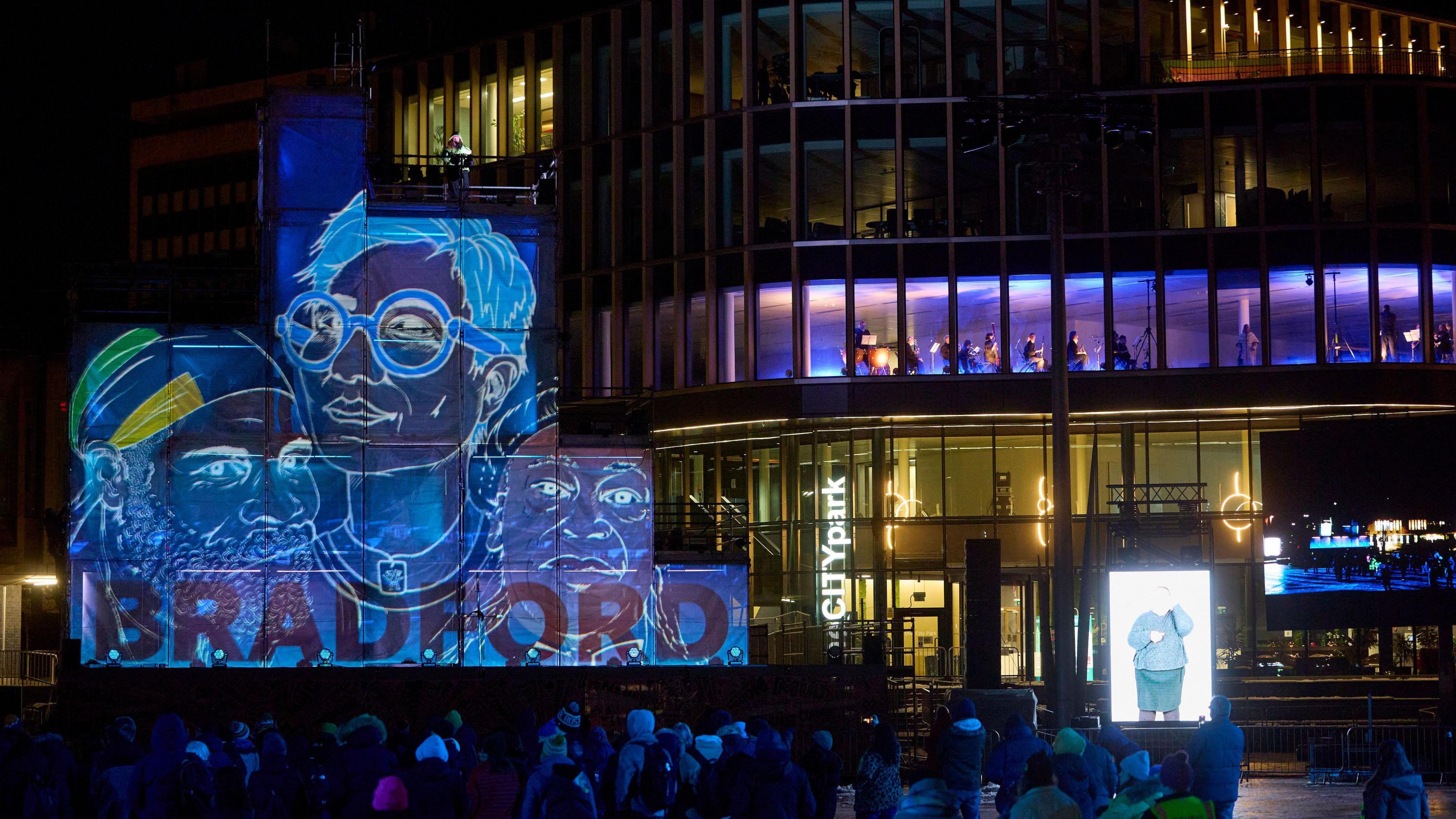 An orchestra are lit up in an office building. In front of them a stage made of blocks has a hand drawn image of people projected onto it. A crowd stands in front.