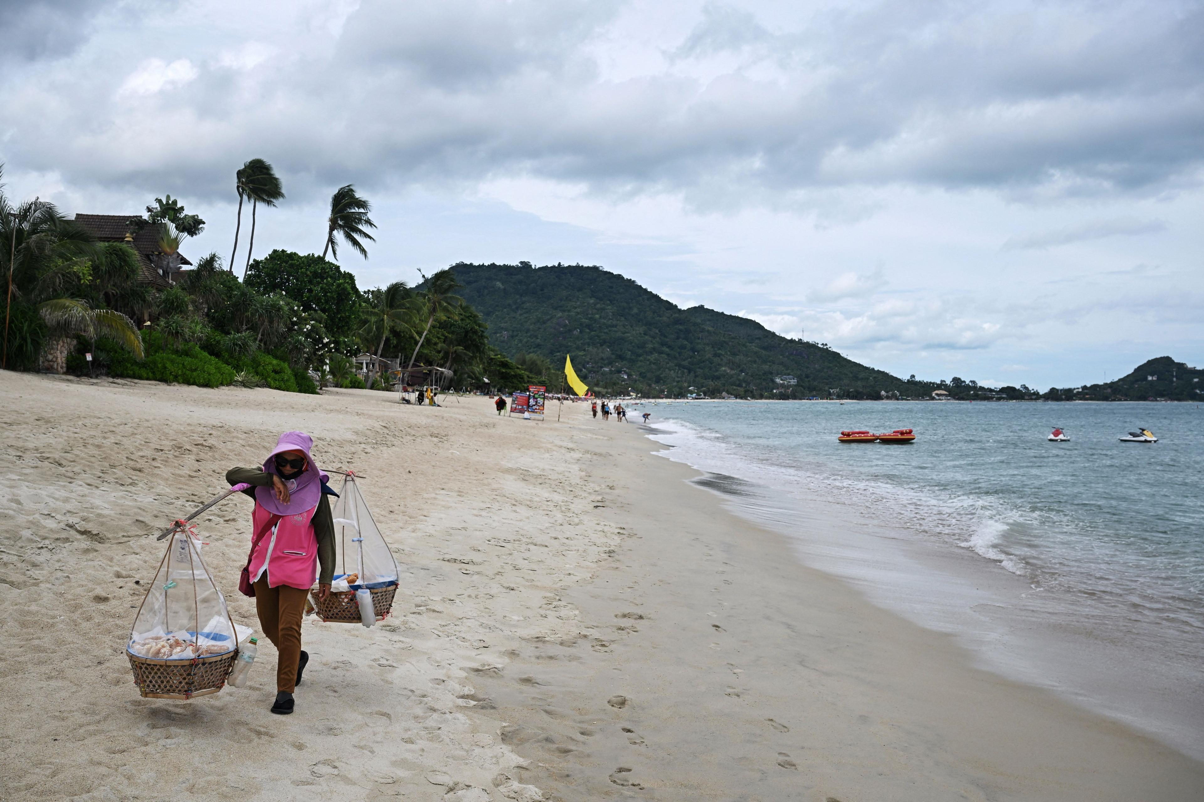 A vendor wearing a purple hat and sunglasses carries donuts on Lamai beach close to the sea on the southern Thai island of Koh Samui