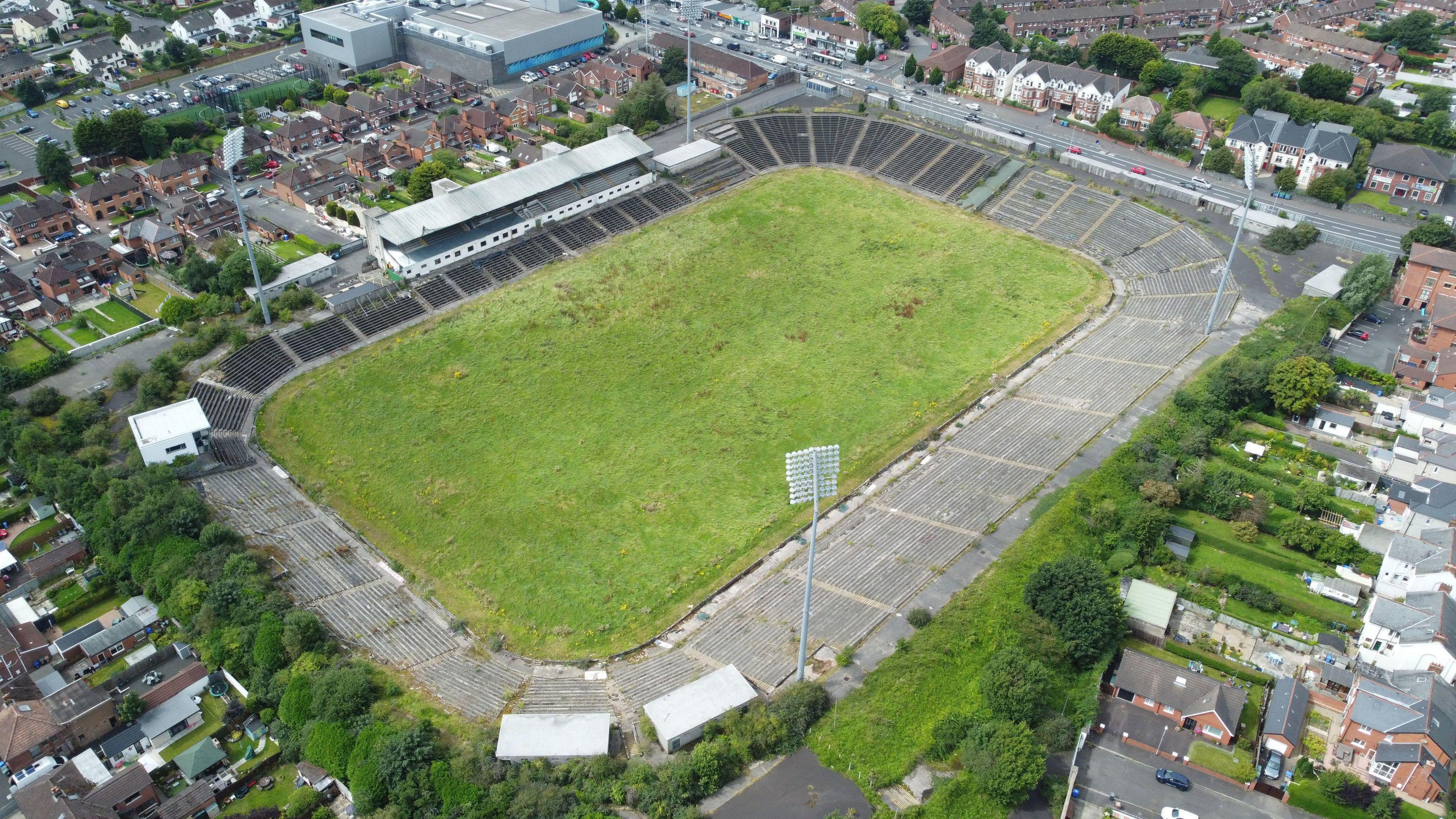 A green grass sports pitch is surrounded by seating. There are metal floodlights around the stadium. Around the exterior of the stadium is houses and a road.