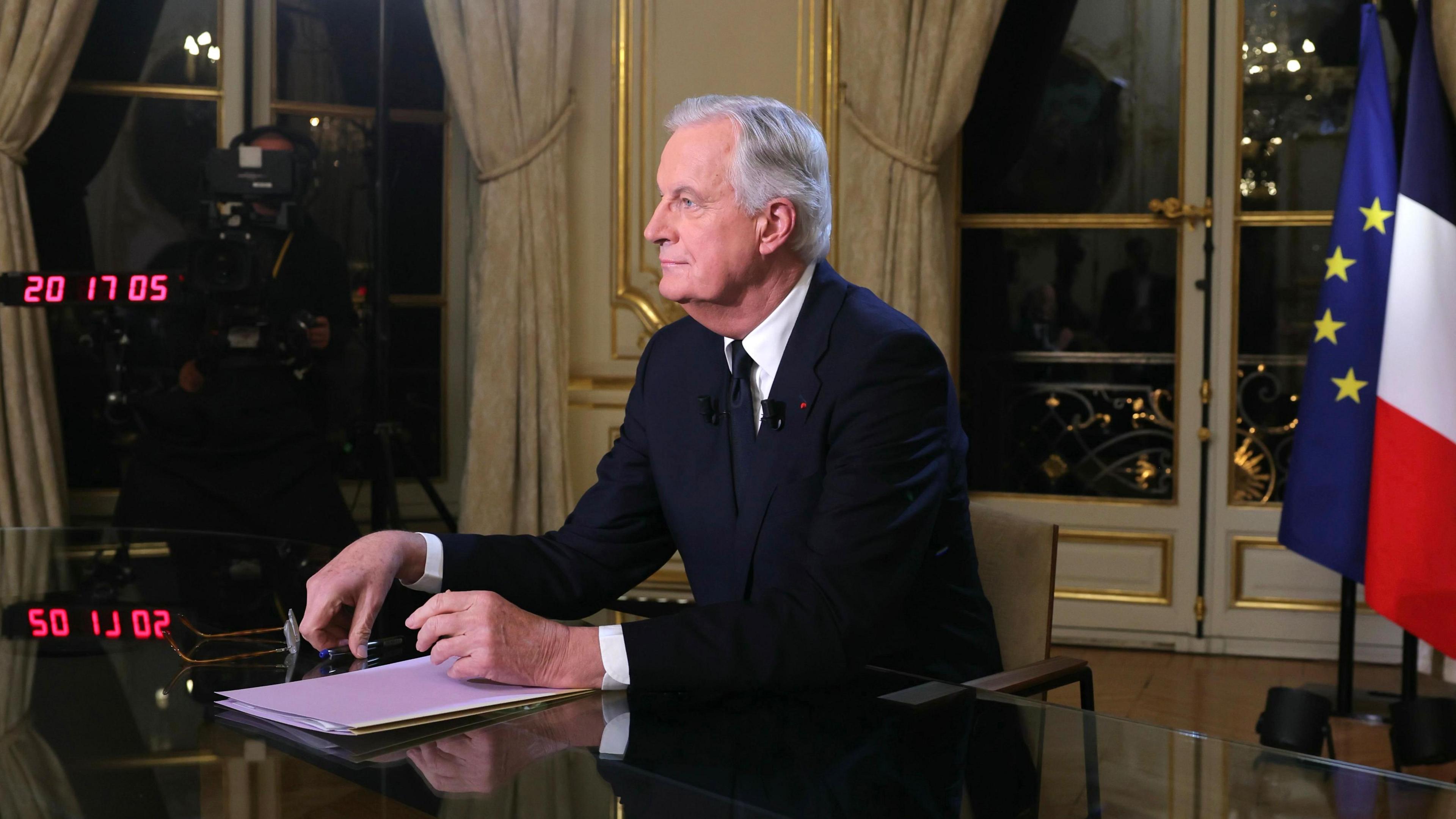 Michel Barnier, who has grey combed over hair and wears a black suit with a white shirt and a black tie, looks towards a camera during a TV interview at the Hotel Matignon in Paris 