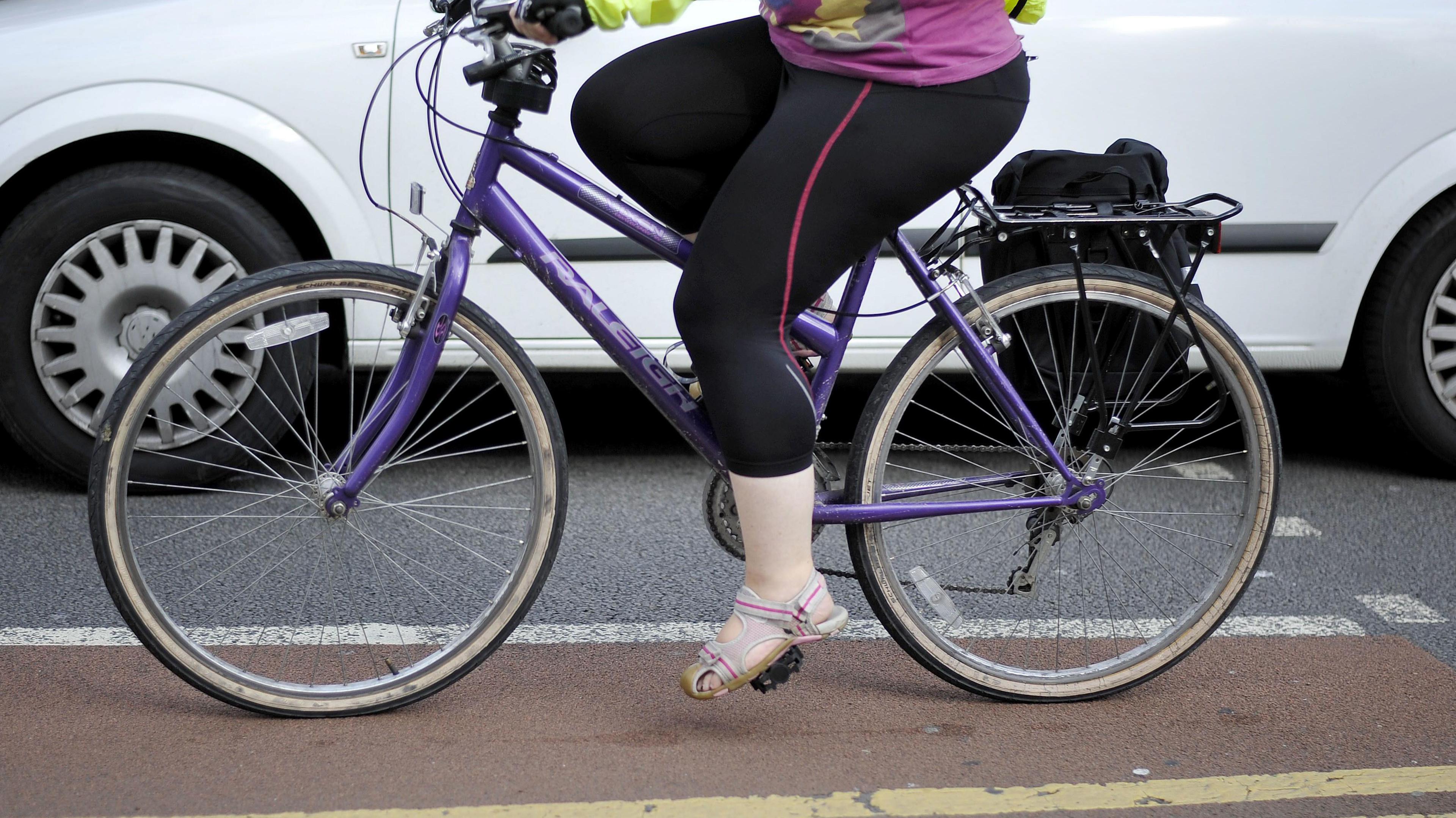 A woman wearing grey open toed sandals, black leggings with a red line on them, a pink t-shirt with a yellow and grey pattern on it and a high-vis jacket cycles on a purple bike in a red cycle lane while a white van drives past her. She has a black bag attached to the rear of her bicycle.