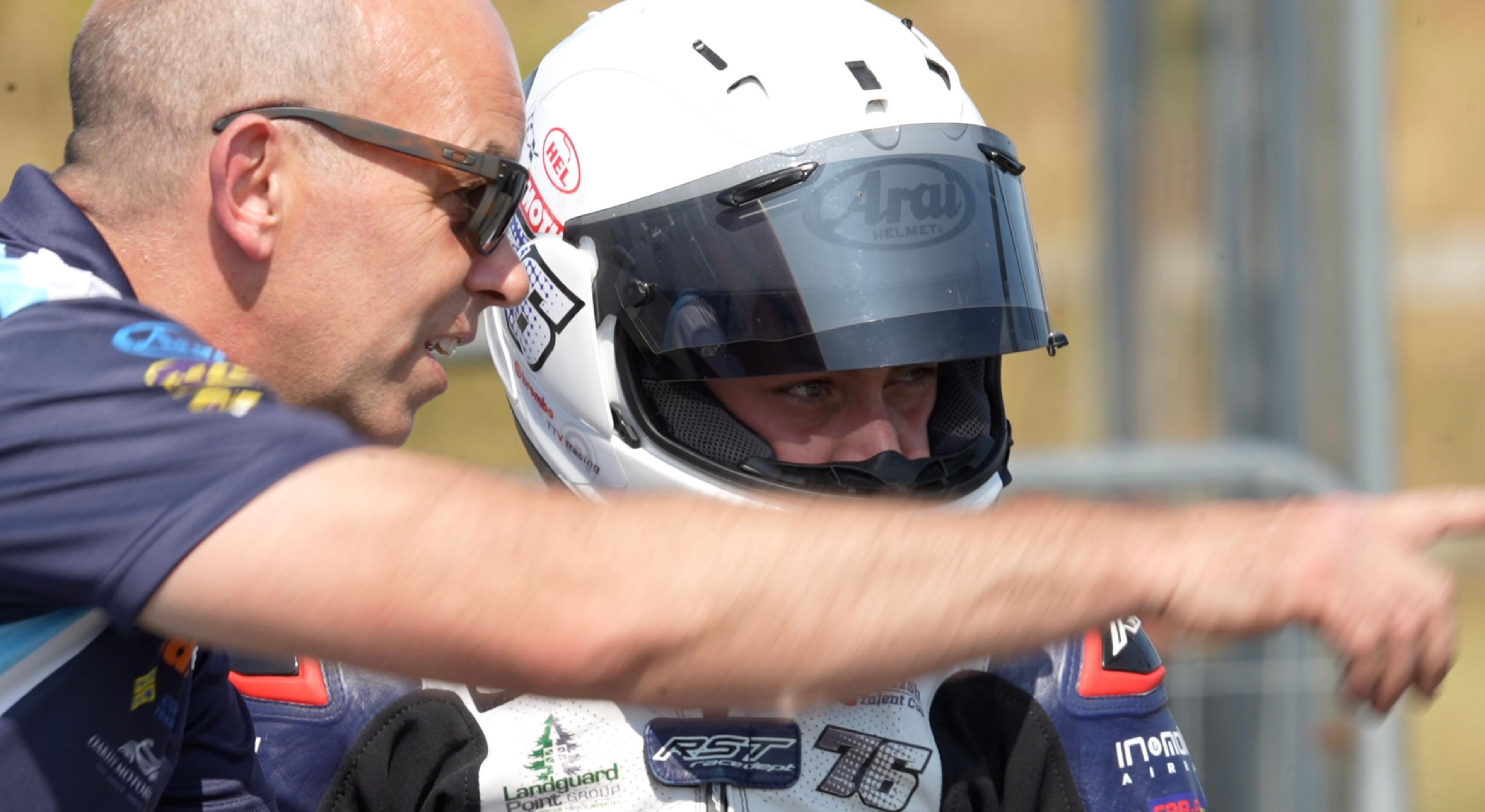 Simon Huntingford, who is bald and wearing sunglasses, is leaning across Charlie and instructing. Charlie is in his full blue leathers, and his white helmet, while sat stationary on his bike, absorbing his manager's information.
