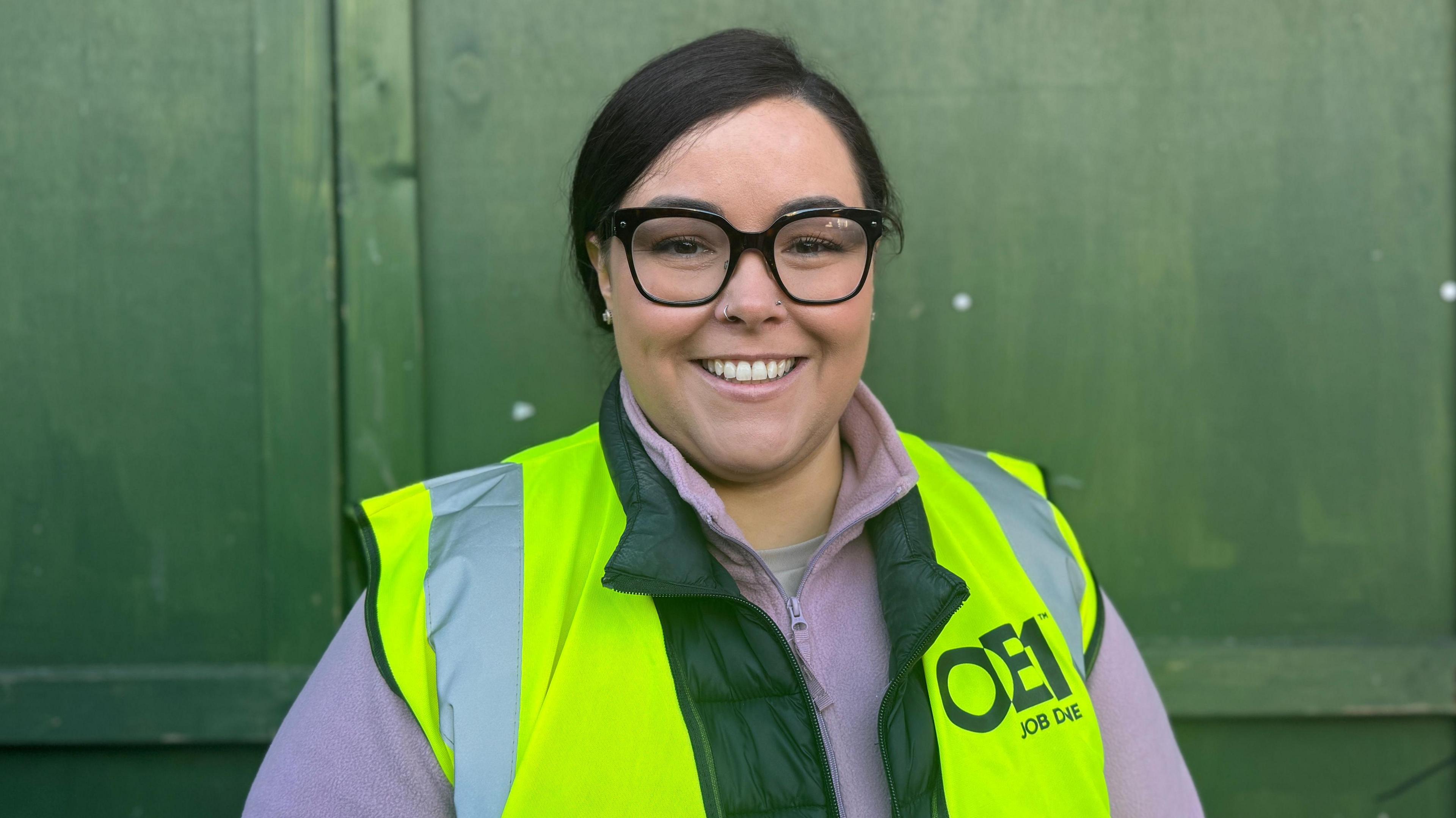 Emma Goodman - a woman with dark hair tied back, wearing black square-rimmed glasses, a lilac zip-up hoodie and a yellow hi-vis jacket smiles at the camera while standing in front of green wooden doors.