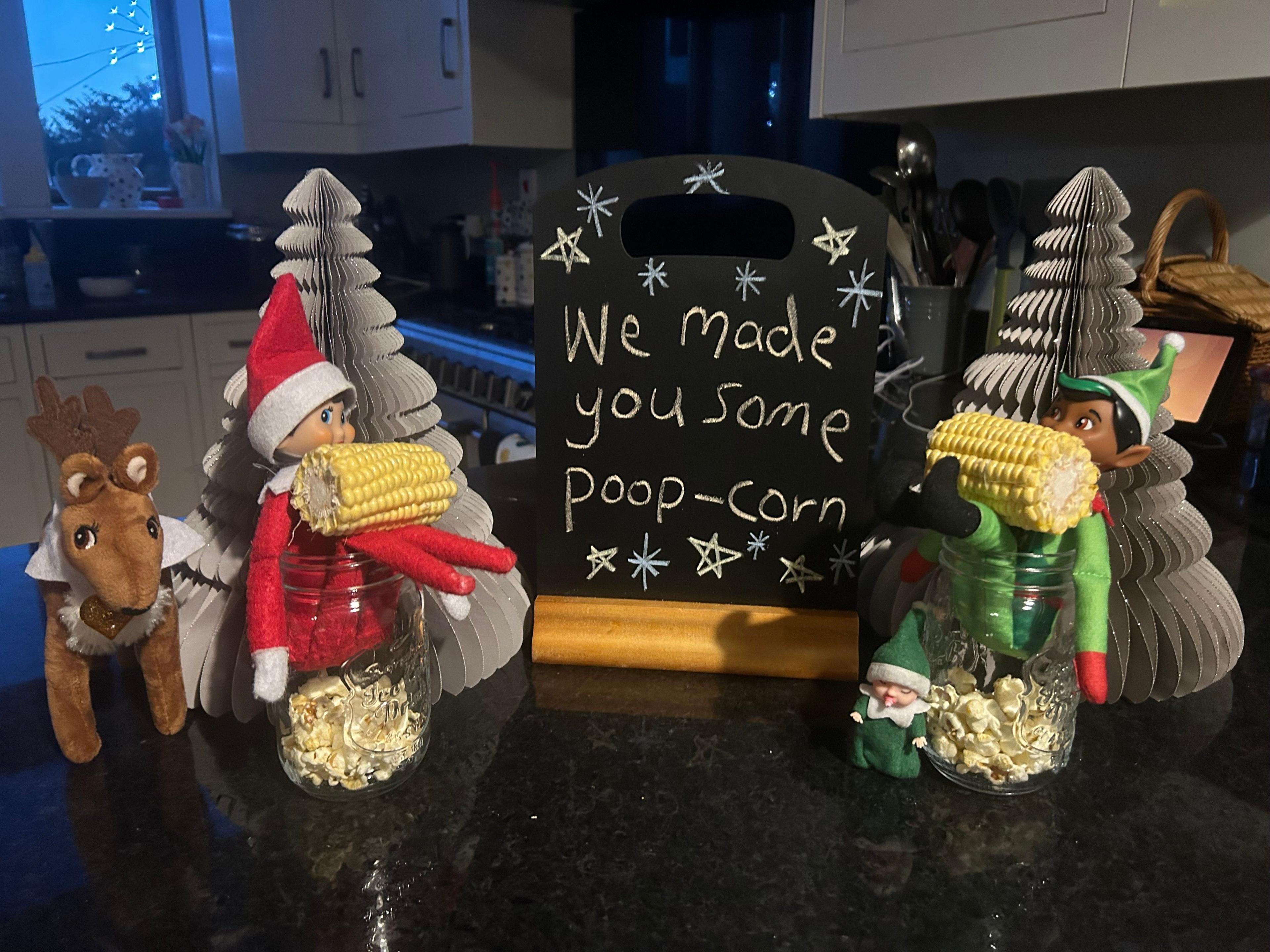 Elves seen sitting inside in a glass jar with popcorn at the bottom. Their legs are sticking out of the jar and a sign next to them reads: "We made you some poop-corn" 