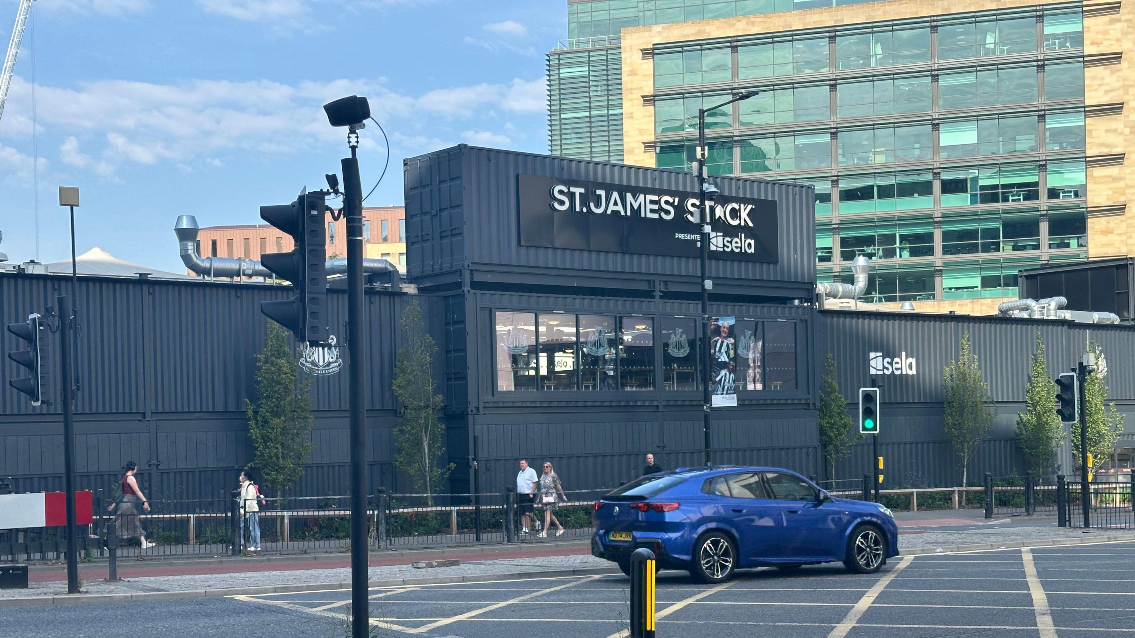 St James' Stack. It is a made up of a number of repurposed shipping containers painted black. A car drives past while a number of people are walking outside.