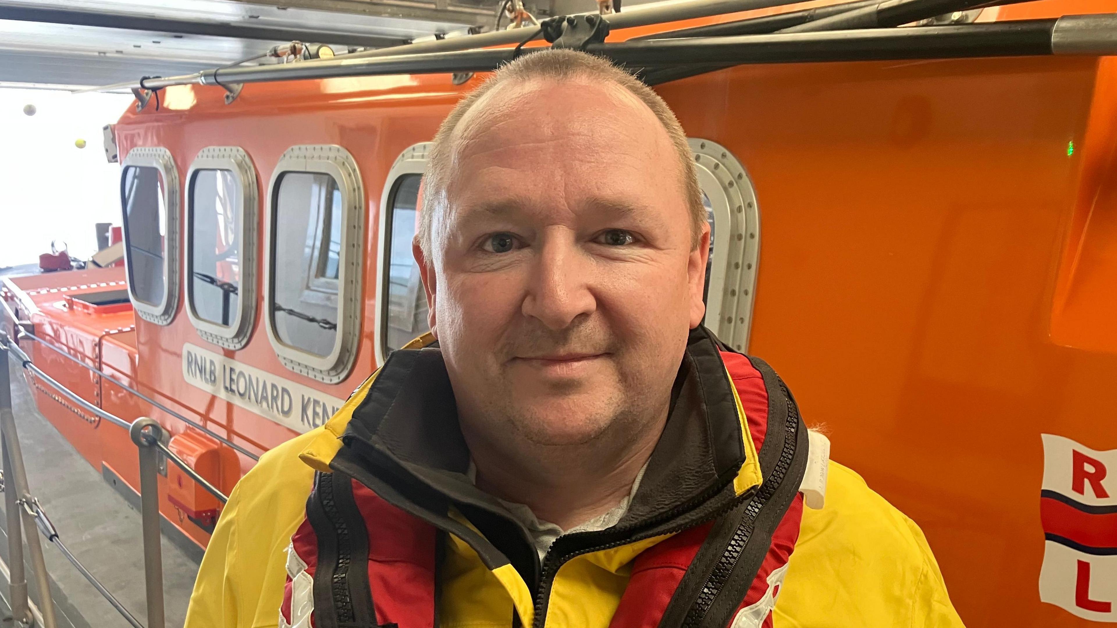 Standing in front of an orange boat is Brendan in his yellow lifeboat crew jacket and red life jacket. He has balding brown hair and brown eyes. He is smiling at the camera.