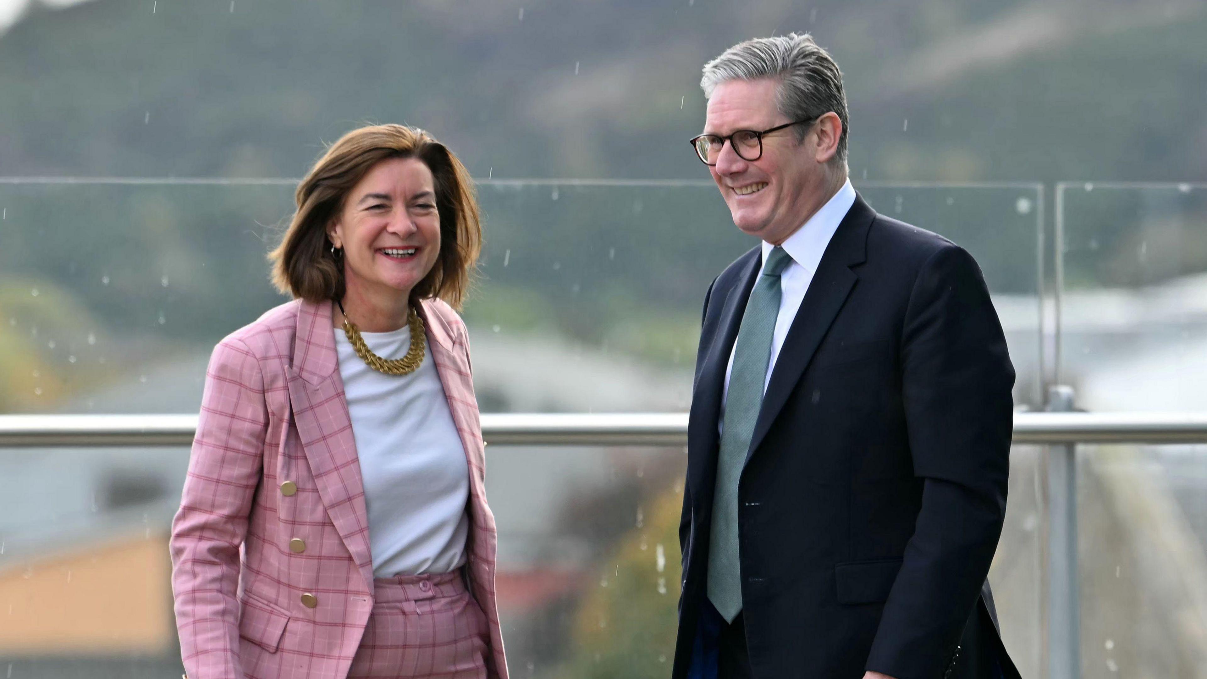 Prime Minister Sir Keir Starmer stands to the right of Wales' First Minister Eluned Morgan. He is wearing a dark suit and grey tie. She is wearing a pink checked trouser suit and necklace