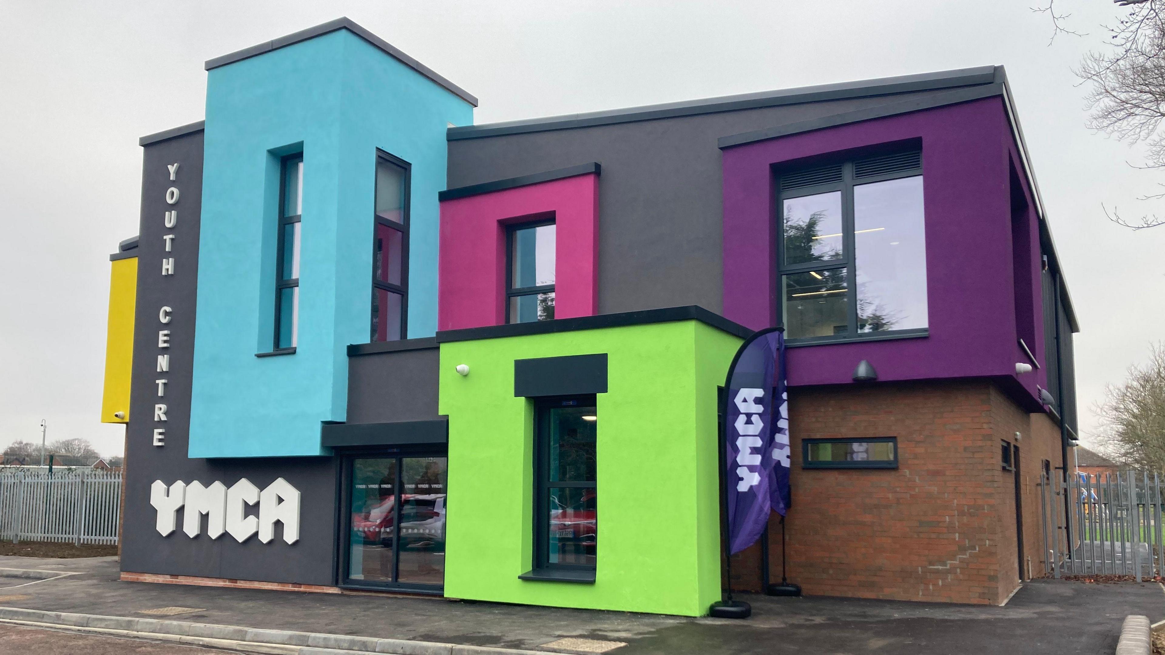 The exterior of the newly refurbished YMCA Youth Centre. It is a brightly-coloured, two-storey, modern building. Sections of the cladding are painted purple, lime green, pink, pale blue and yellow. Running down one side of the building are the words "Youth Centre". Lower down is written "YMCA"