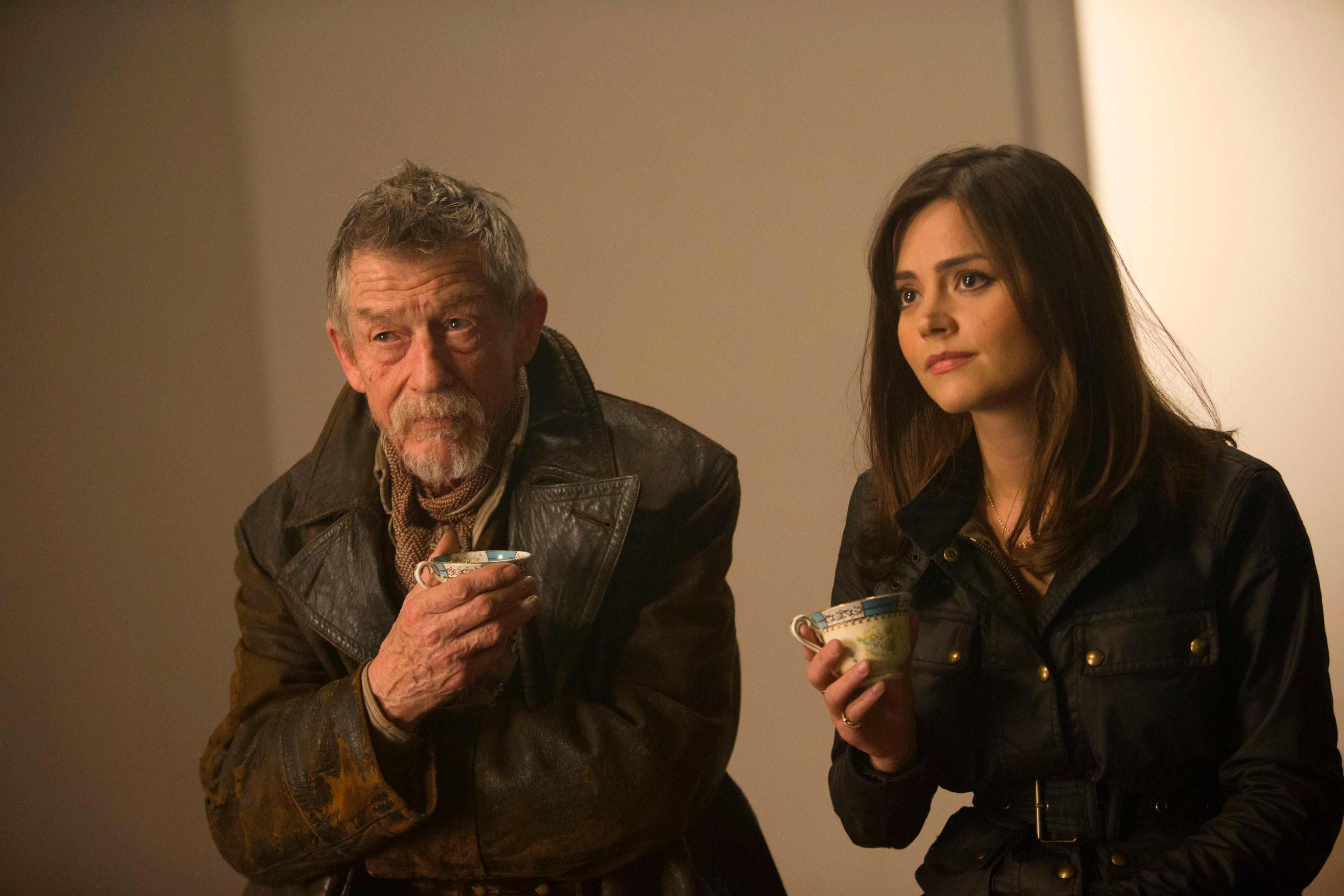 The War Doctor, who has short greying hair and a white goatee beard and is wearing a battered brown leather coat with a scarf around his neck, is sitting next to Clara, who has long dark hair and is wearing a black, belted jacket. They both hold an old fashioned small teacup in their right hands.