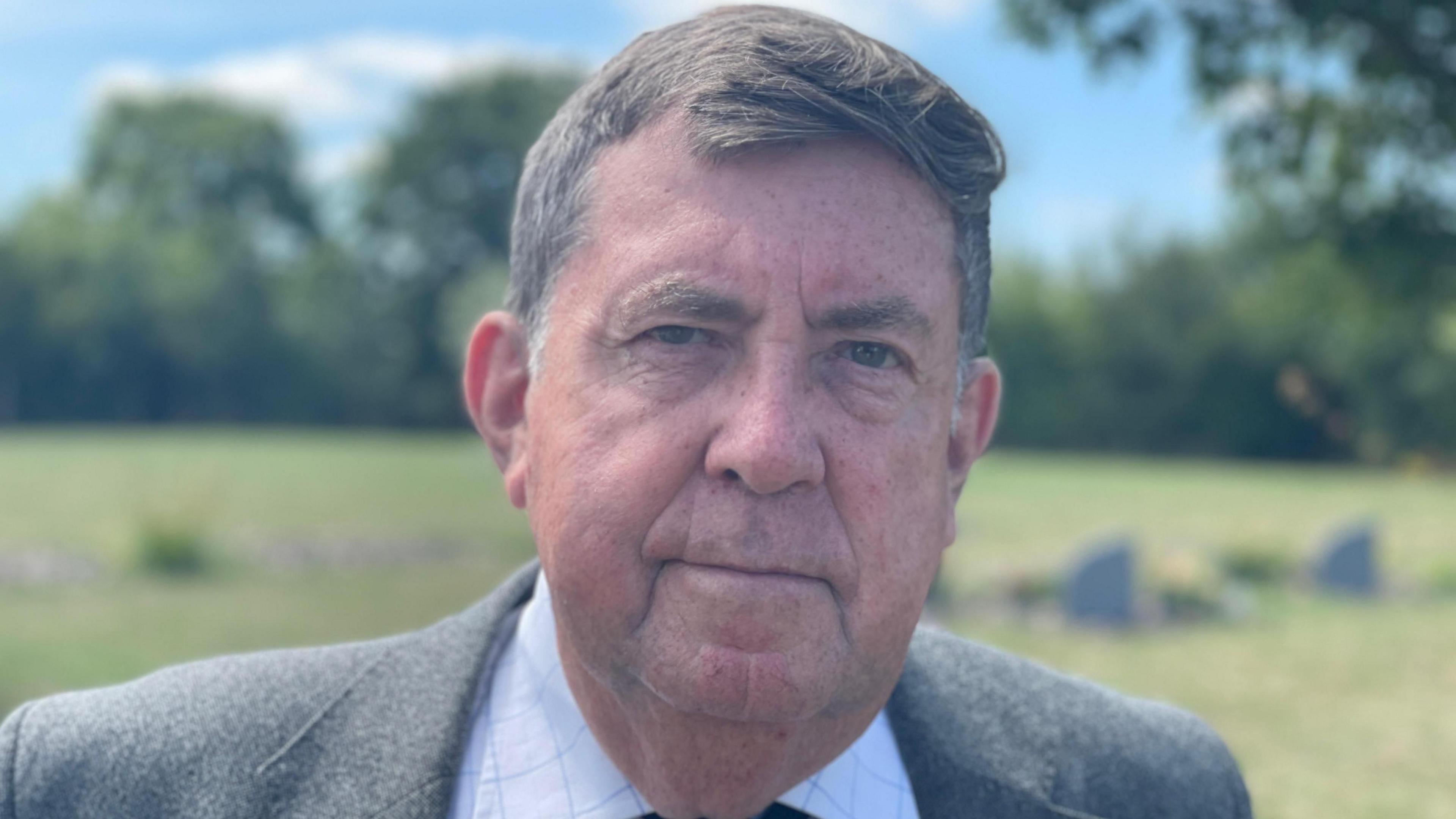 Richard Crozier is looking towards the camera with a serious look. He has short grey hair and is wearing a white shirt and grey suit jacket. He is standing on a field.