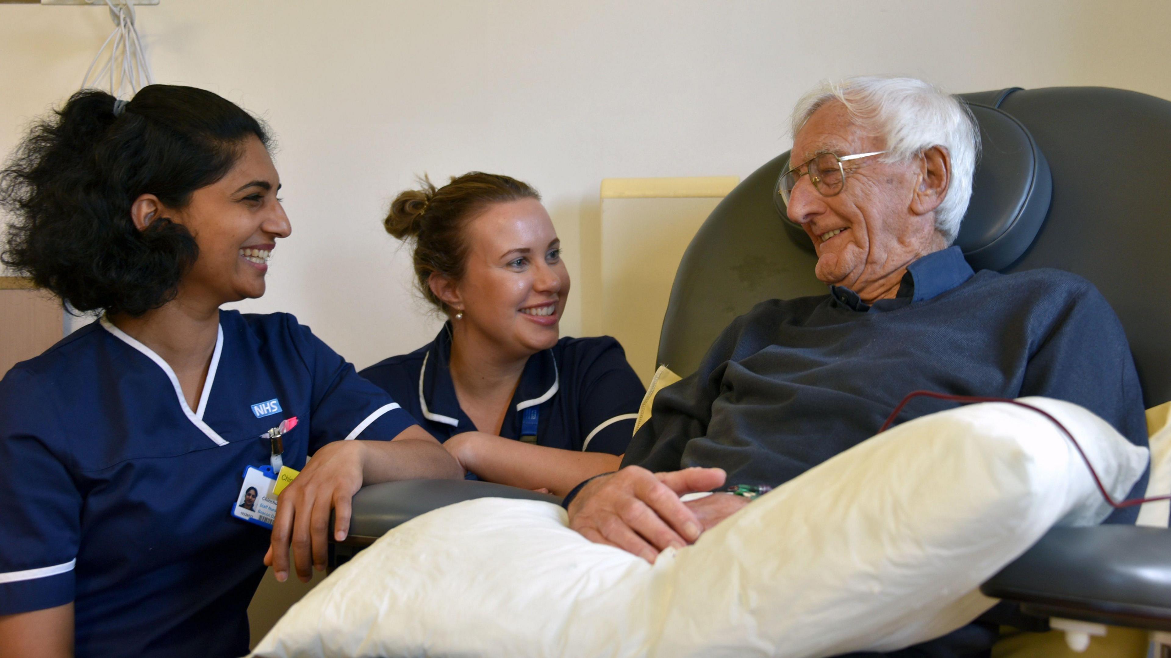 Left to right is Chinchu Abraham and Beth Quick - staff nurses, and Andre Guerrier - patient. Chinchu and Beth are wearing NHS blue scrubs and are leaning on the chair which Andre is sat in. Andre is wearing a blue jumper and has a pillow across his lap.