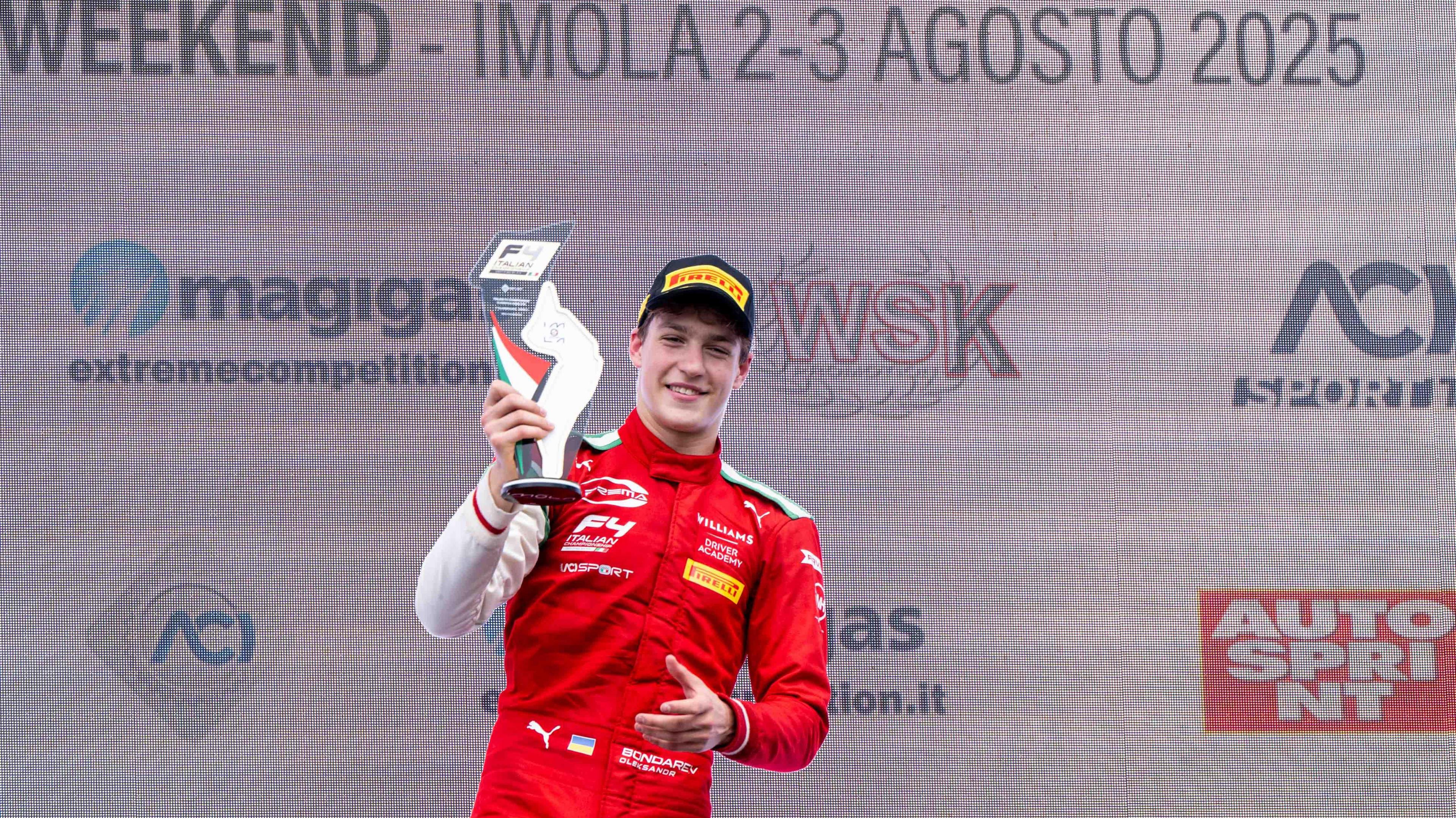 Bondarev dressed up in his red racing kit lifting up the winning trophy from his victory at Imola. He is smiling and looking at the camera. The backdrop displays the various sponsors of the event.