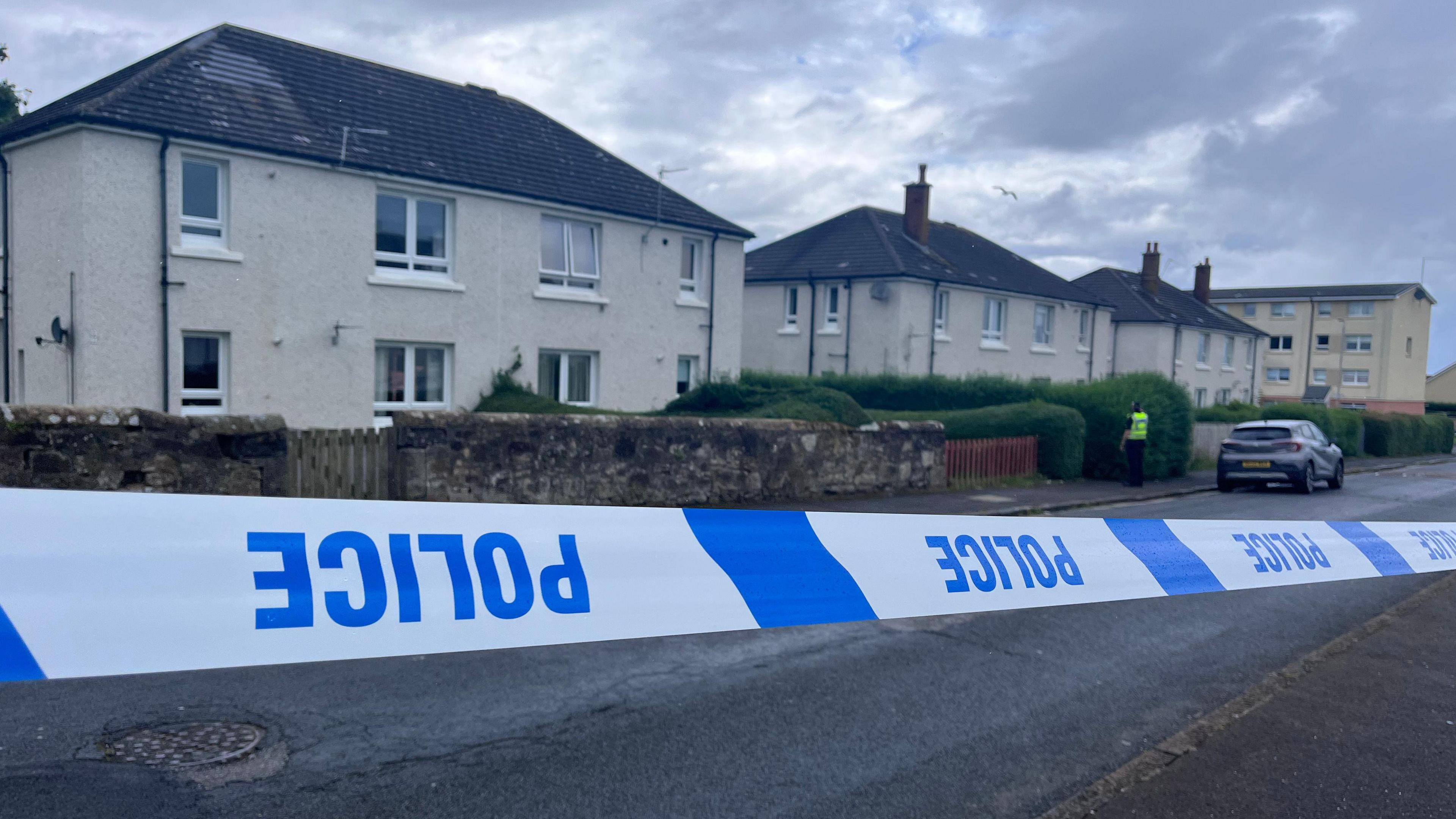 
Blue and white police tape which is upside down in front of a row of cream two-storey buildings. A police officer is visible in a distance.