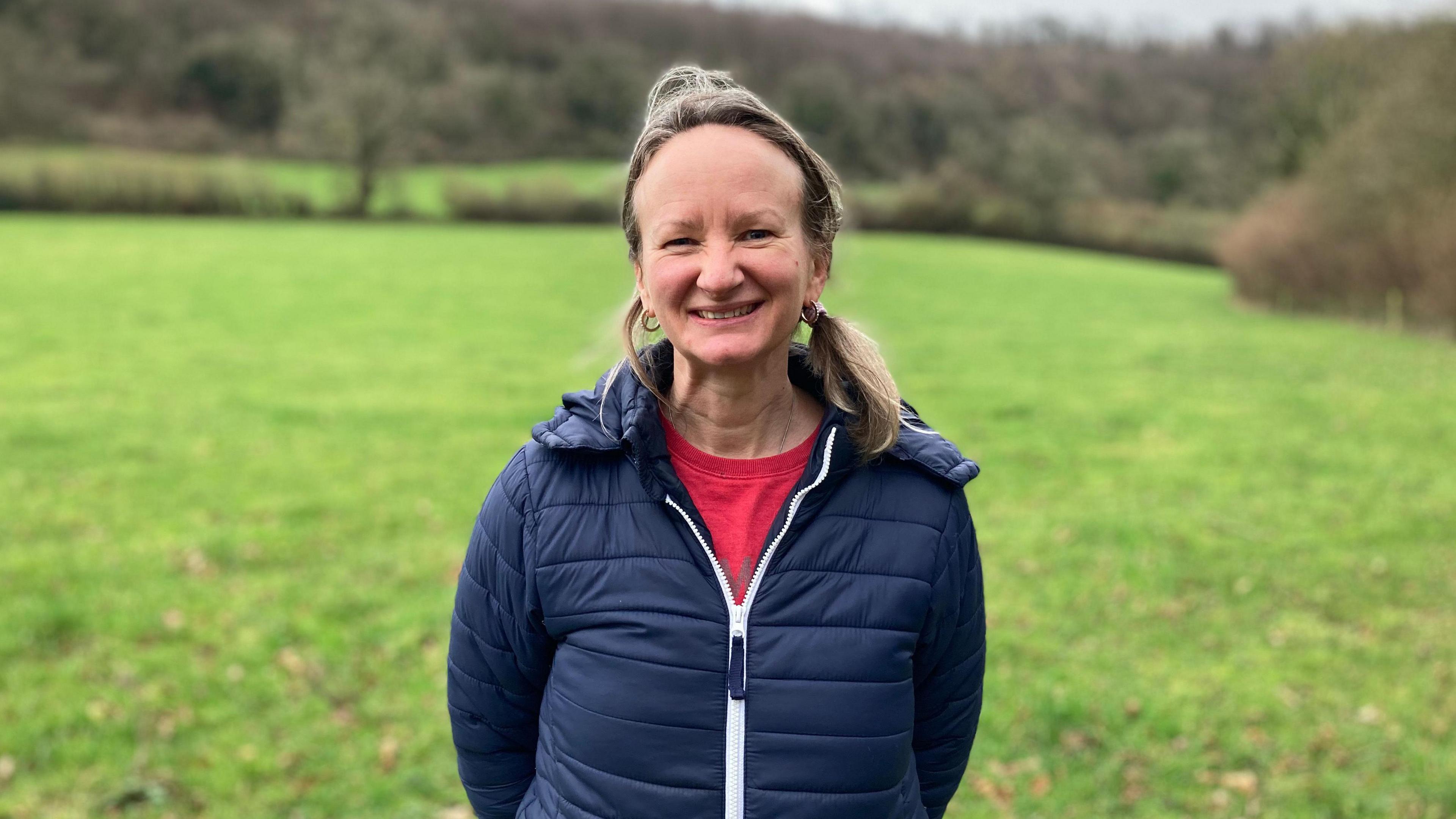 Amy Morris wearing a pink t-shirt underneath a blue padded zip up jacket. She has her blonde hair tied back in a ponytail on her left shoulder, and is smiling at the camera. She is standing in a field on Court Farm where the pools could be built. 