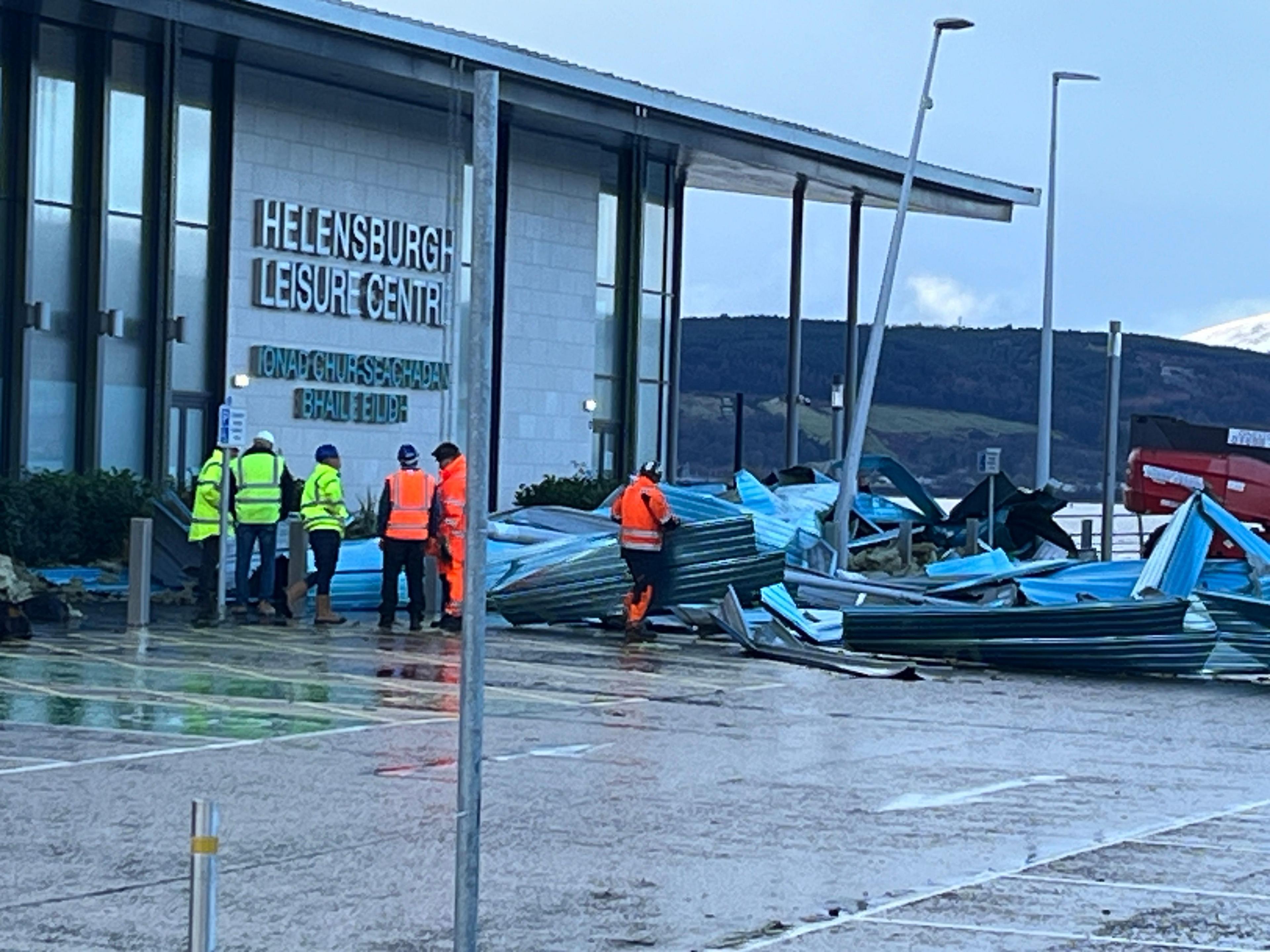 A group of six men in high visibility yellow and orange jackets are working to clear a large amount of metal debris from the car park of the Helensburgh leisure centre. A street lamp is leaning over at an alarming angle.