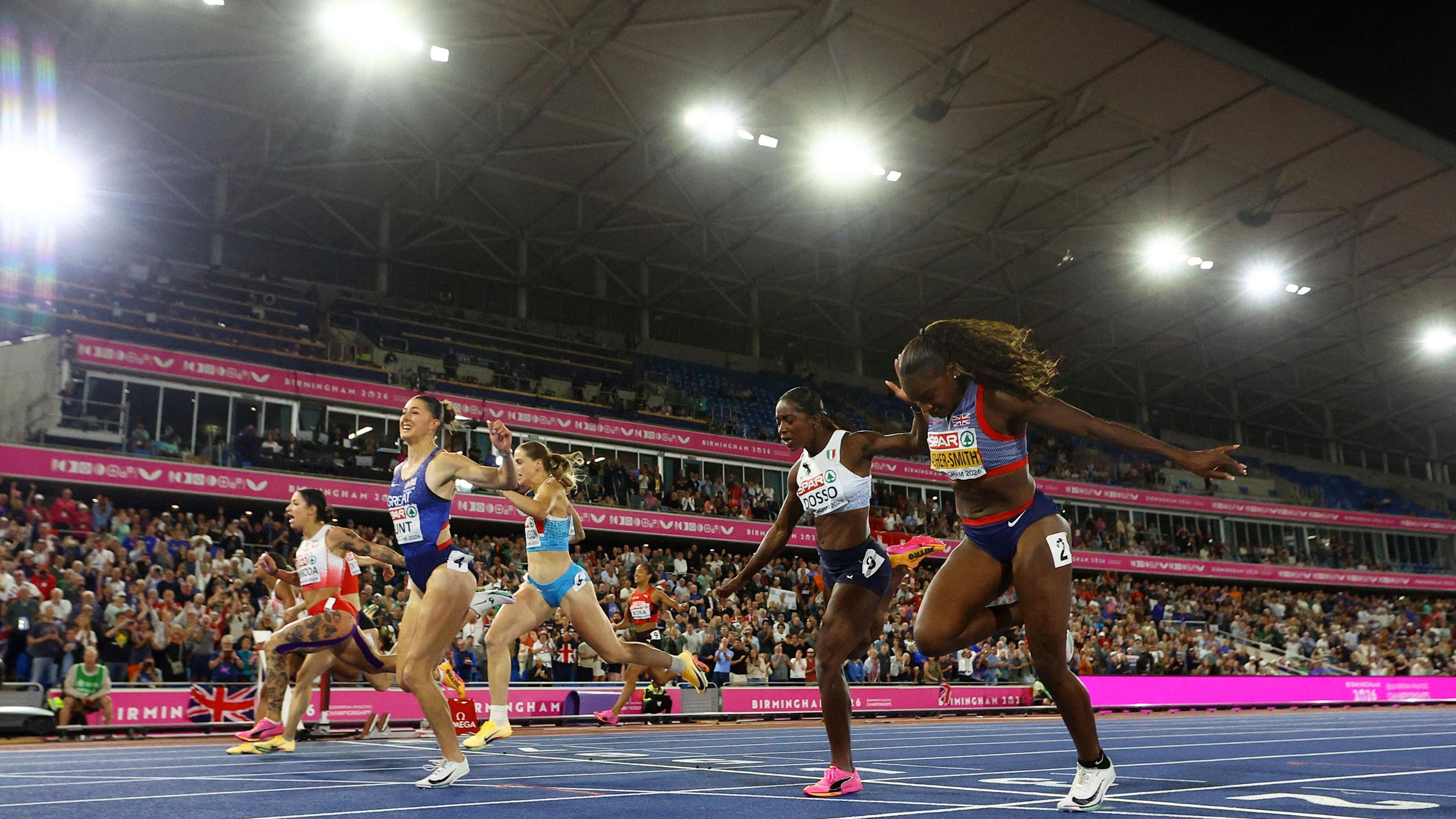 A photo of six women in a race. The floor is blue and the stands are behind which are not full. The signs are pink. 