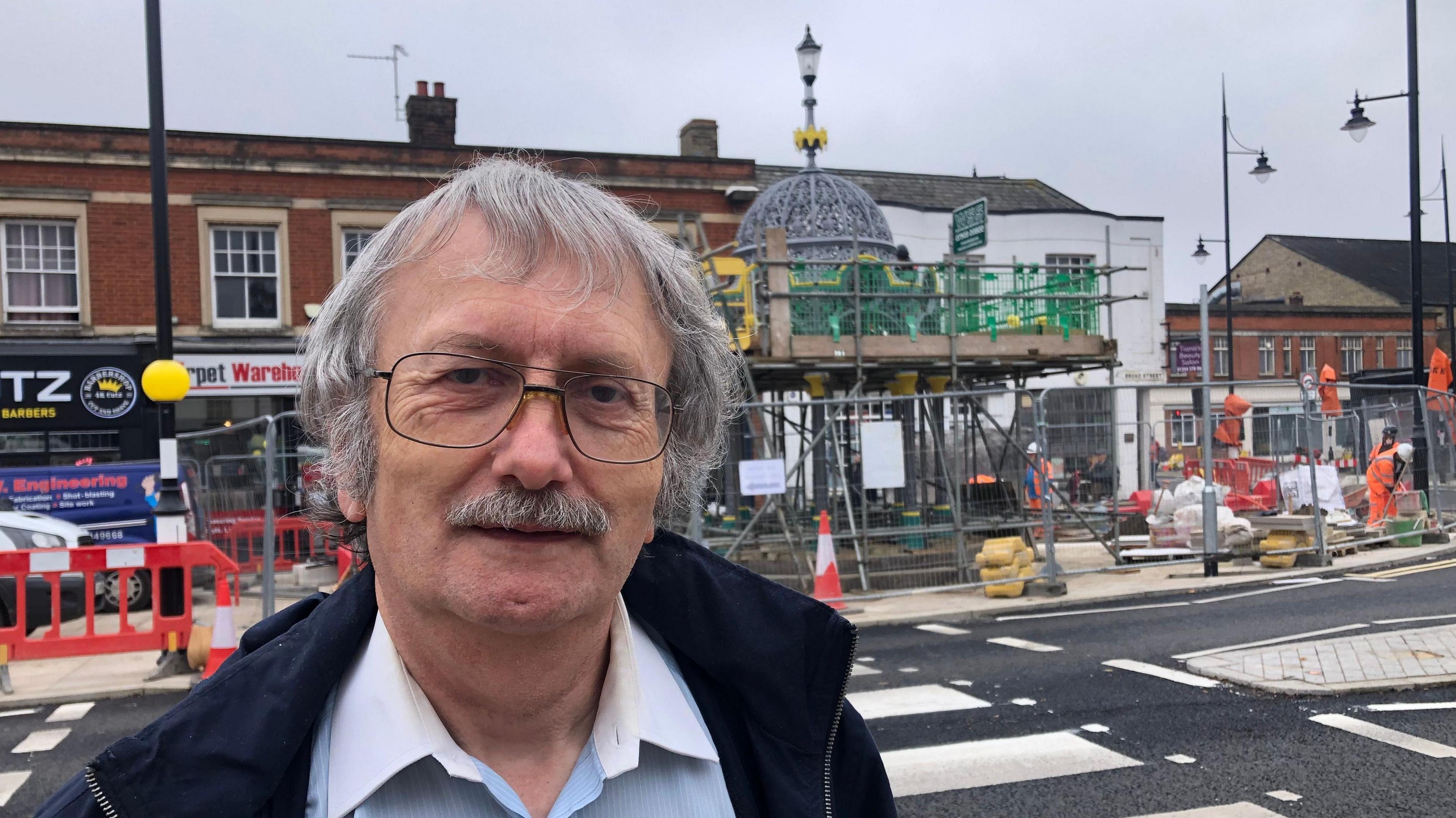 David Edwards has light grey hair, a grey moustache and glasses, he is wearing a dark blue fleece over a light blue shirt with white collar open. Behind him are some workmen in high-vis orange outfits working on the Fountain, which is surrounded by scaffolding.