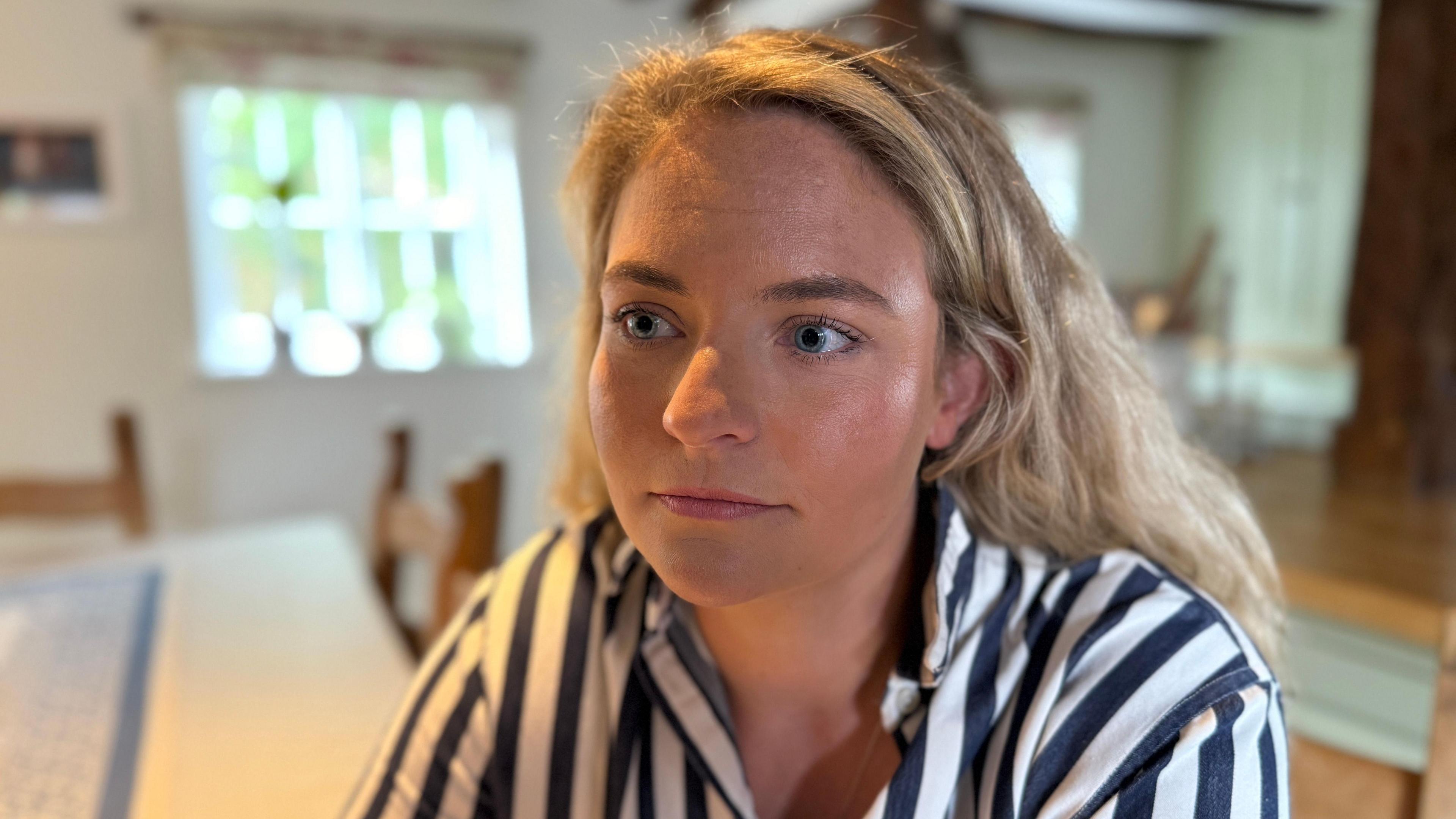 Hannah is looking off to the left of the screen. She has blonde wavy hair that is pushed back behind her ears. She is wearing a blue and white striped shirt. Behind her the background is blurred. You can see the dining room table that has a blue runner down the middle. There is a window behind her and a blurred picture of her dog Betsy on the wall.