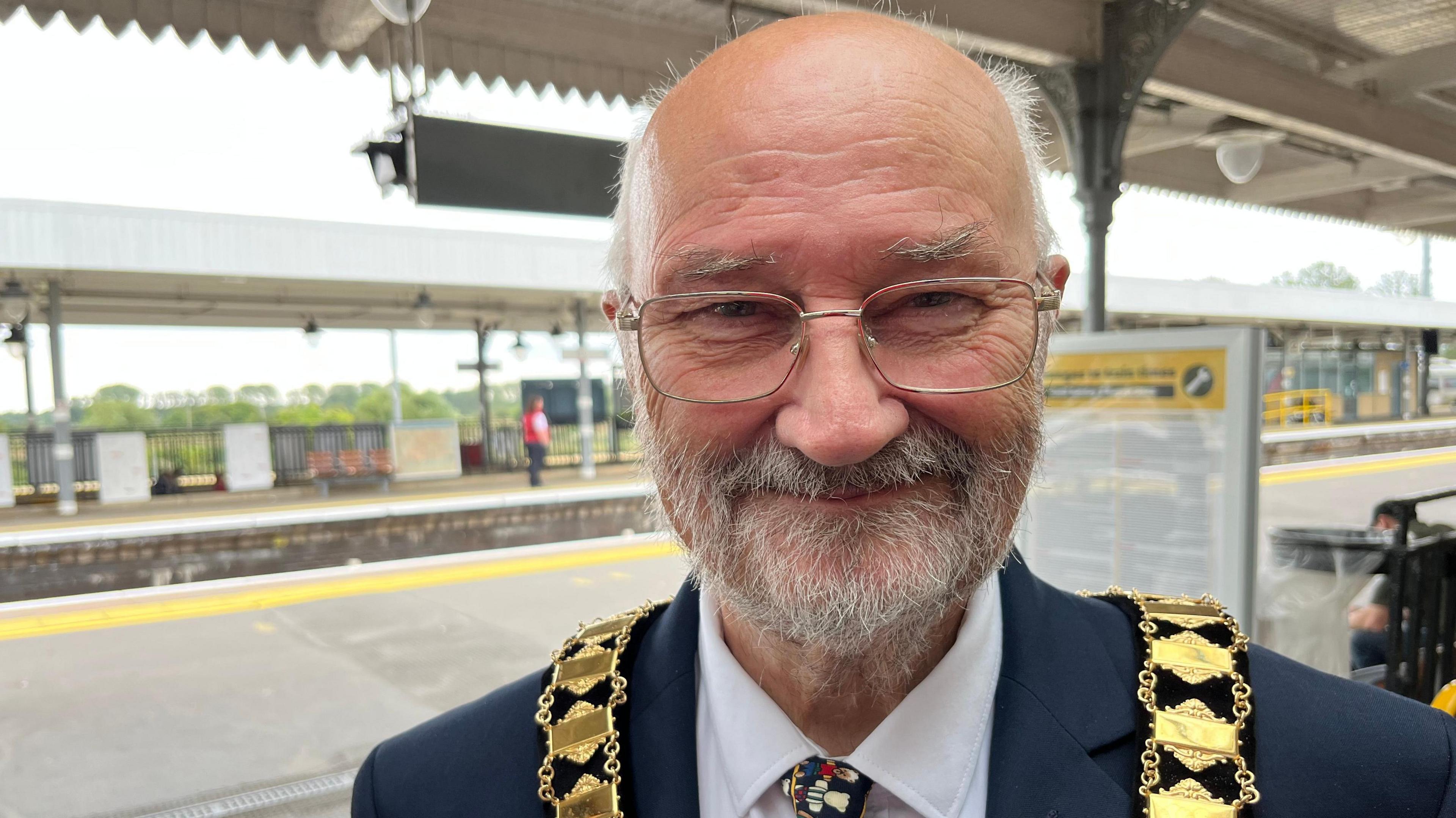 Mike Banyard, with a gold ceremonial chain around his neck, grey hair on the sides of his head, bald on top with a grey beard and moustache and wearing gold rimmed glasses with a blue jacket, white shirt and white, red and blue tie. Behind him is a railway platform.