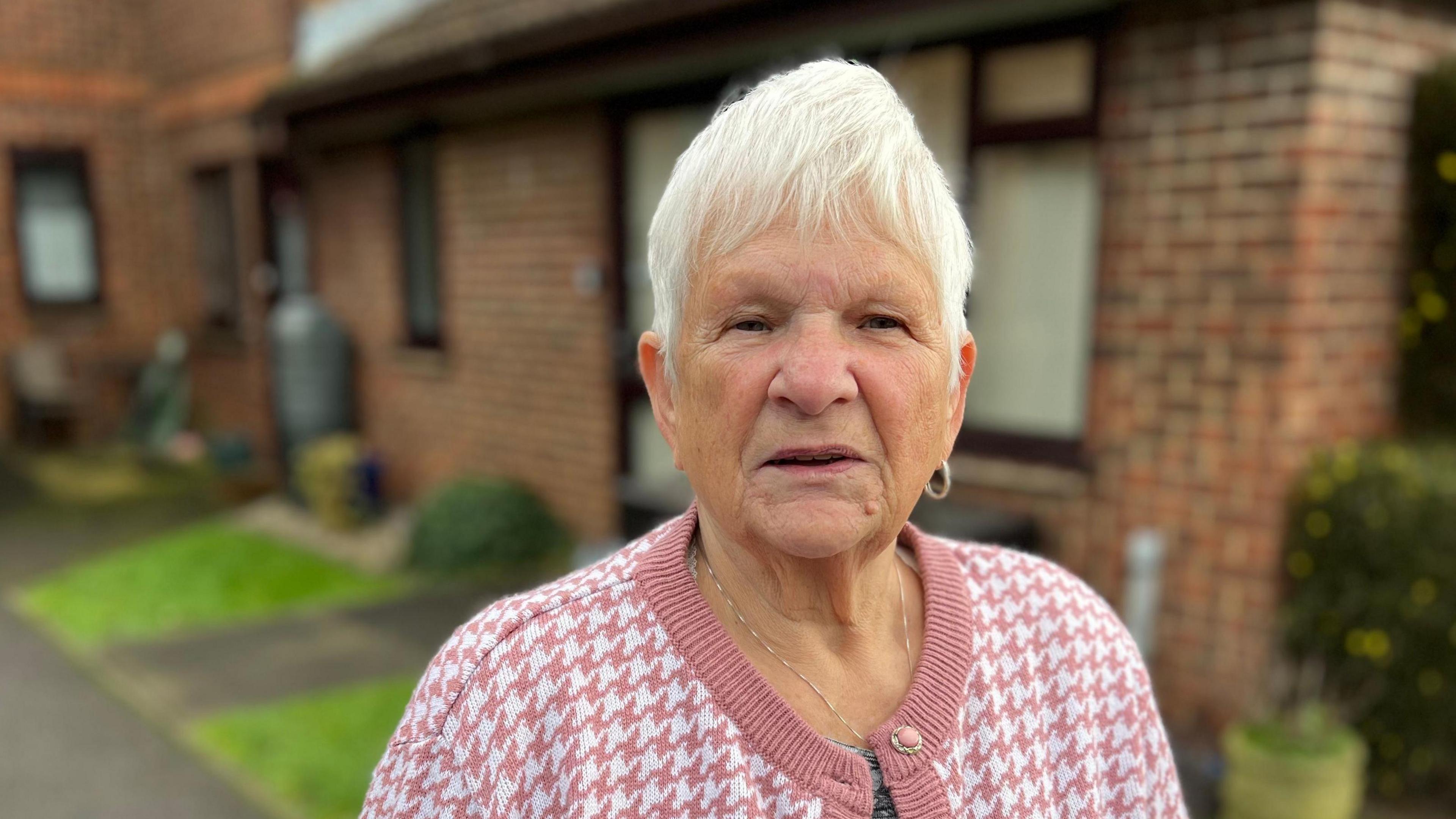 A lady with cropped white hair wearing a pink houndstooth cardigan looks at the camera. A brick house is in the background.