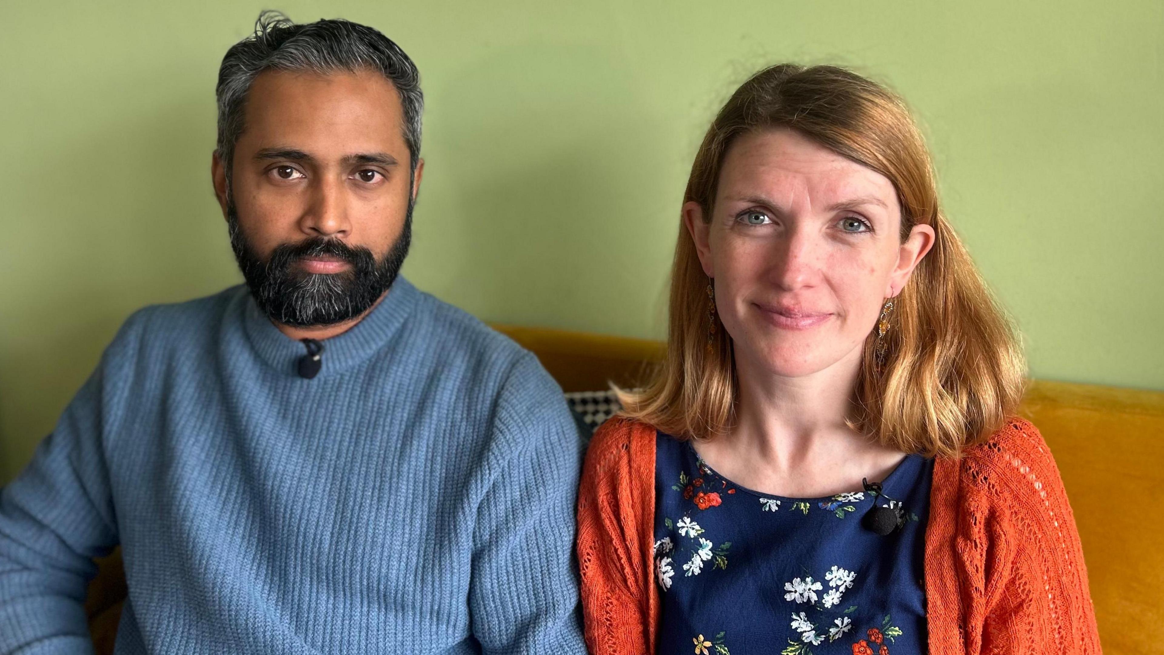 A man, Sree Kadalayil, is on the left and a woman, Jude Pollock, is on the right. They are sitting on an orange sofa that is placed up against a light green wall. Sree has short greying black hair and a black beard with specks of grey. He is wearing a blue jumper. Jude has shoulder-length reddish-blonde hair and is wearing a blue top with a flower pattern and an orange cardigan. 