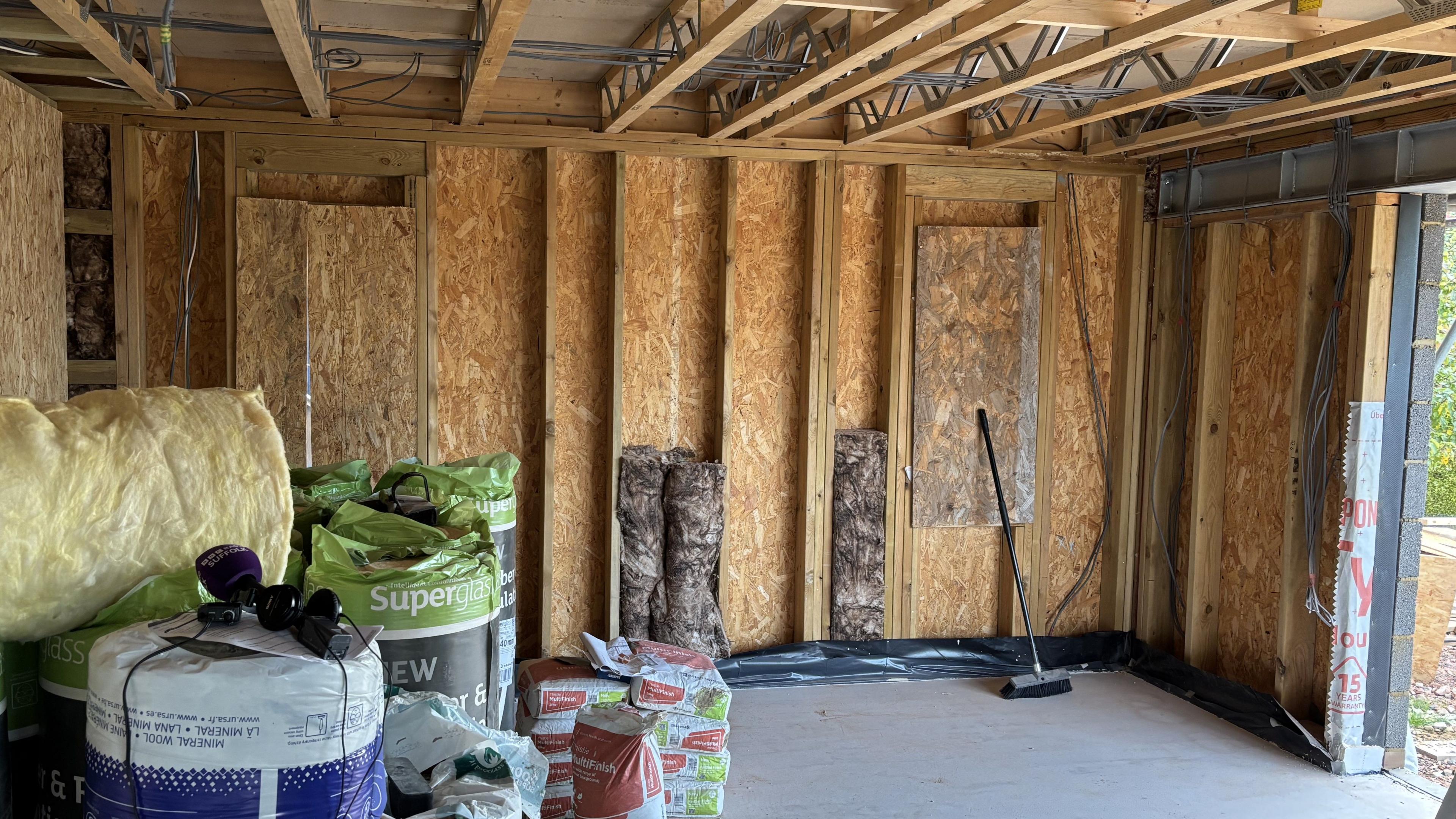 Inside a house that is being renovated. The walls have not yet been plastered and instead wooden beams and panels are on show. House construction materials sit within the room to the left including large rolls of insulation. 