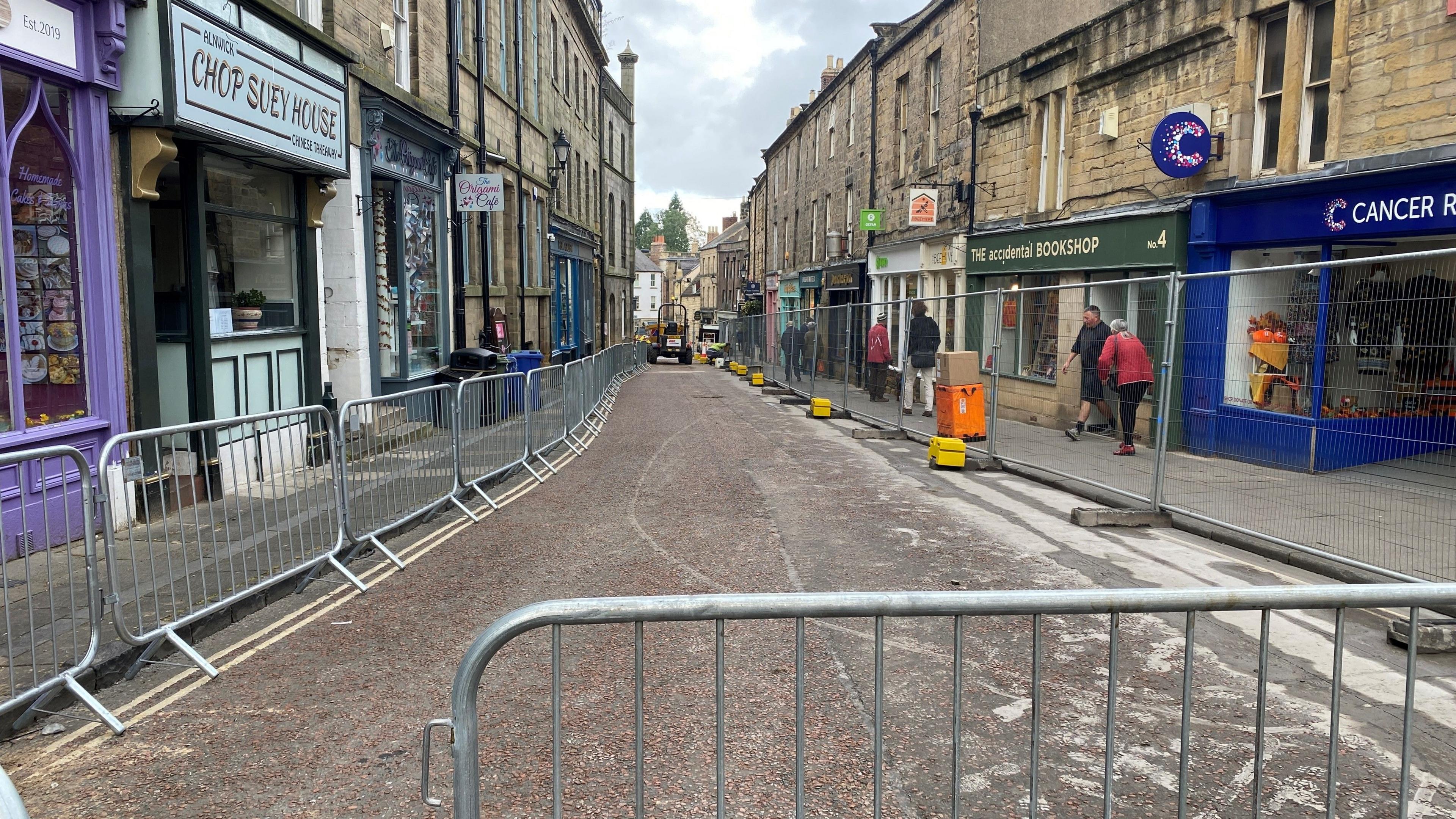 A narrow eighteenth century street has metal fencing along it and a dumper truck can be seen in the distance 