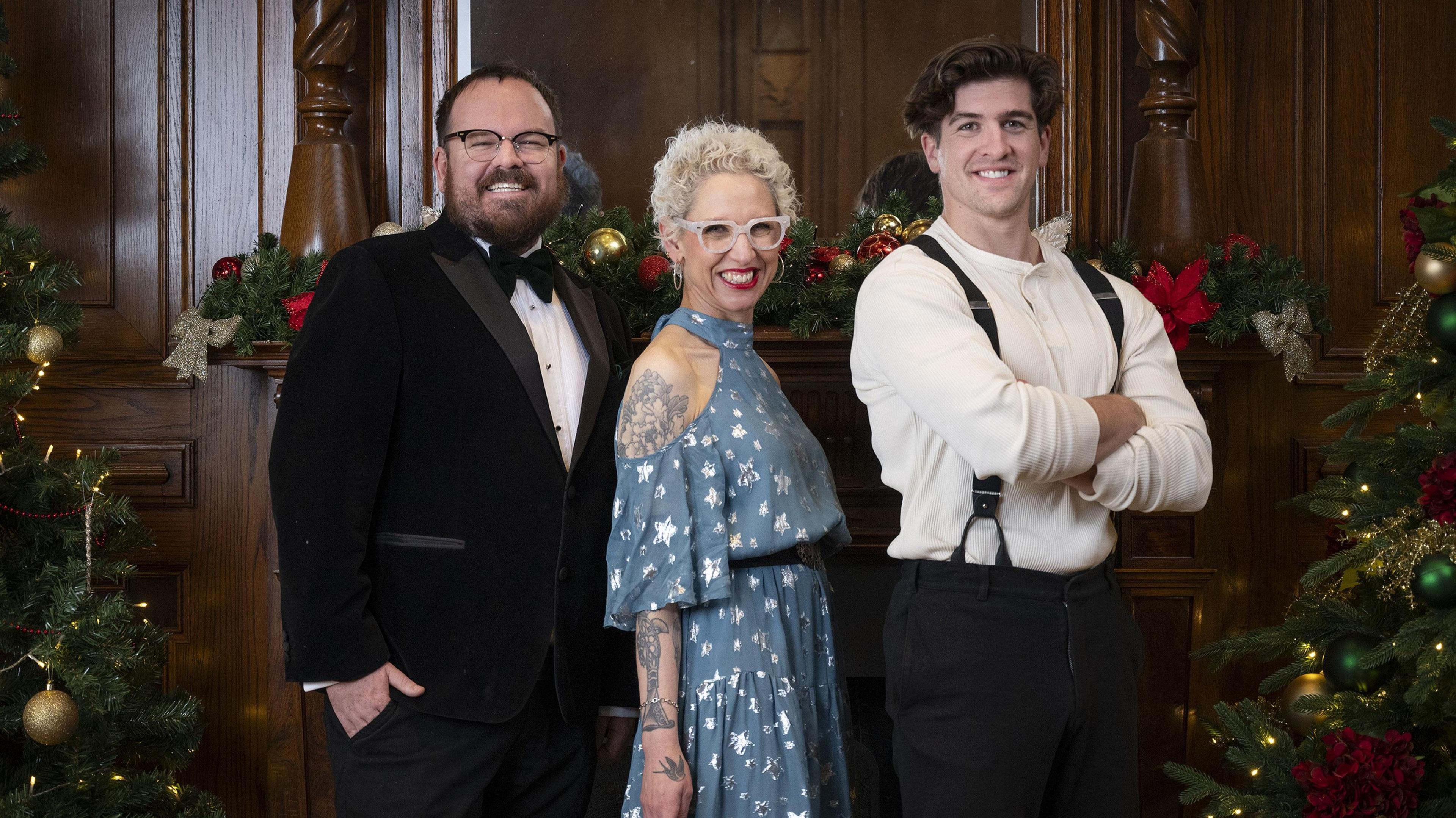 Three people, Anna, Banjo and Danny, stand in a row in front of a wood panelled wall. A Christmas tree stands at either side of them. They all beam happily into the camera.