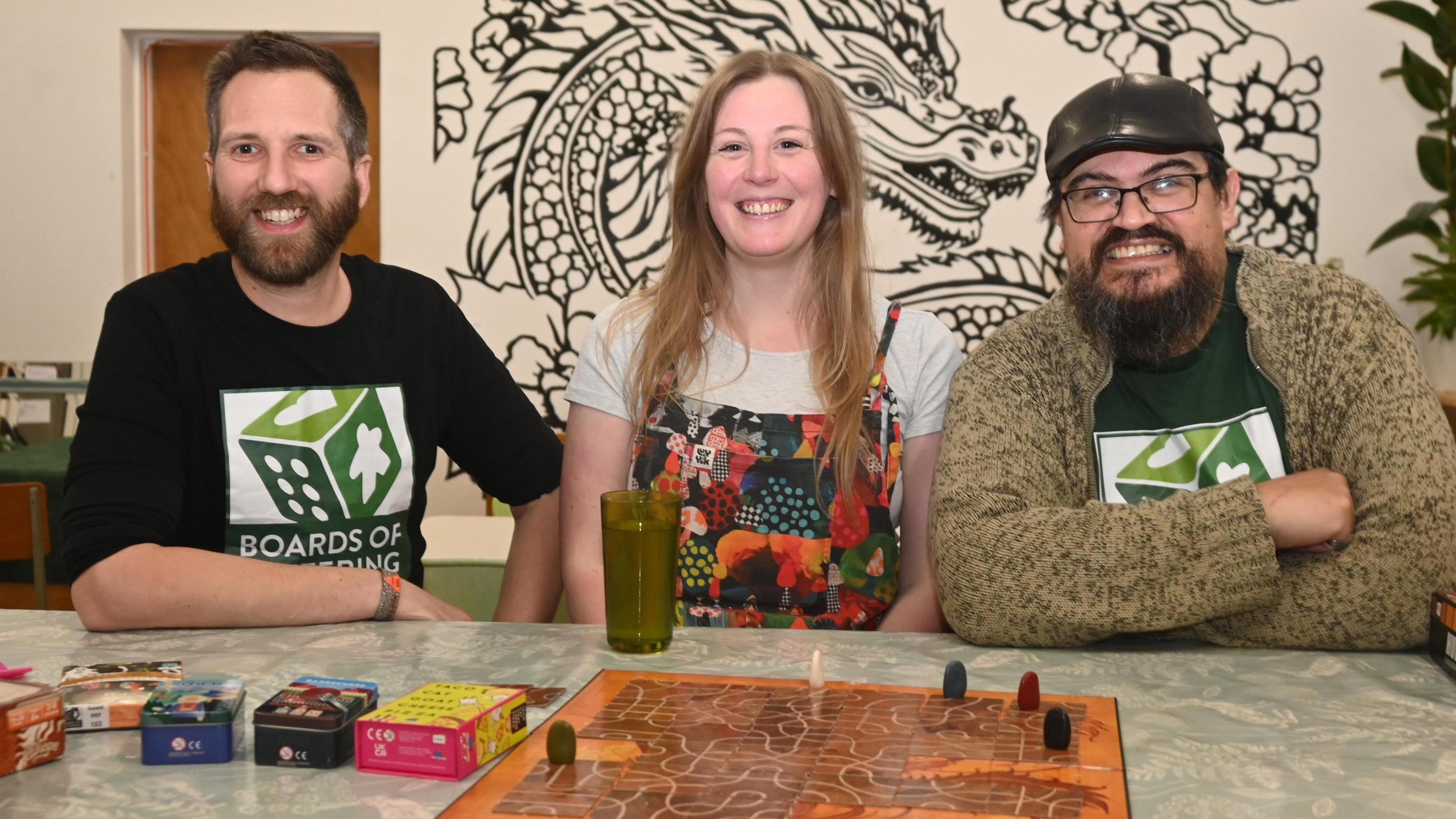 Dez Dell wearing a black tshirt with Boards of Kettering written on it, and a logo of a green and white dice, Polly Shackleton in a white t-shirt and colourful dungarees and Gavin Price in the same t-shirt as Dez Dell and a brown cardigan and black leather cap. Both men have beards and all three are smiling. They are sitting at a table, with all three facing the camera, and a playing board and pieces laid out in front of them.