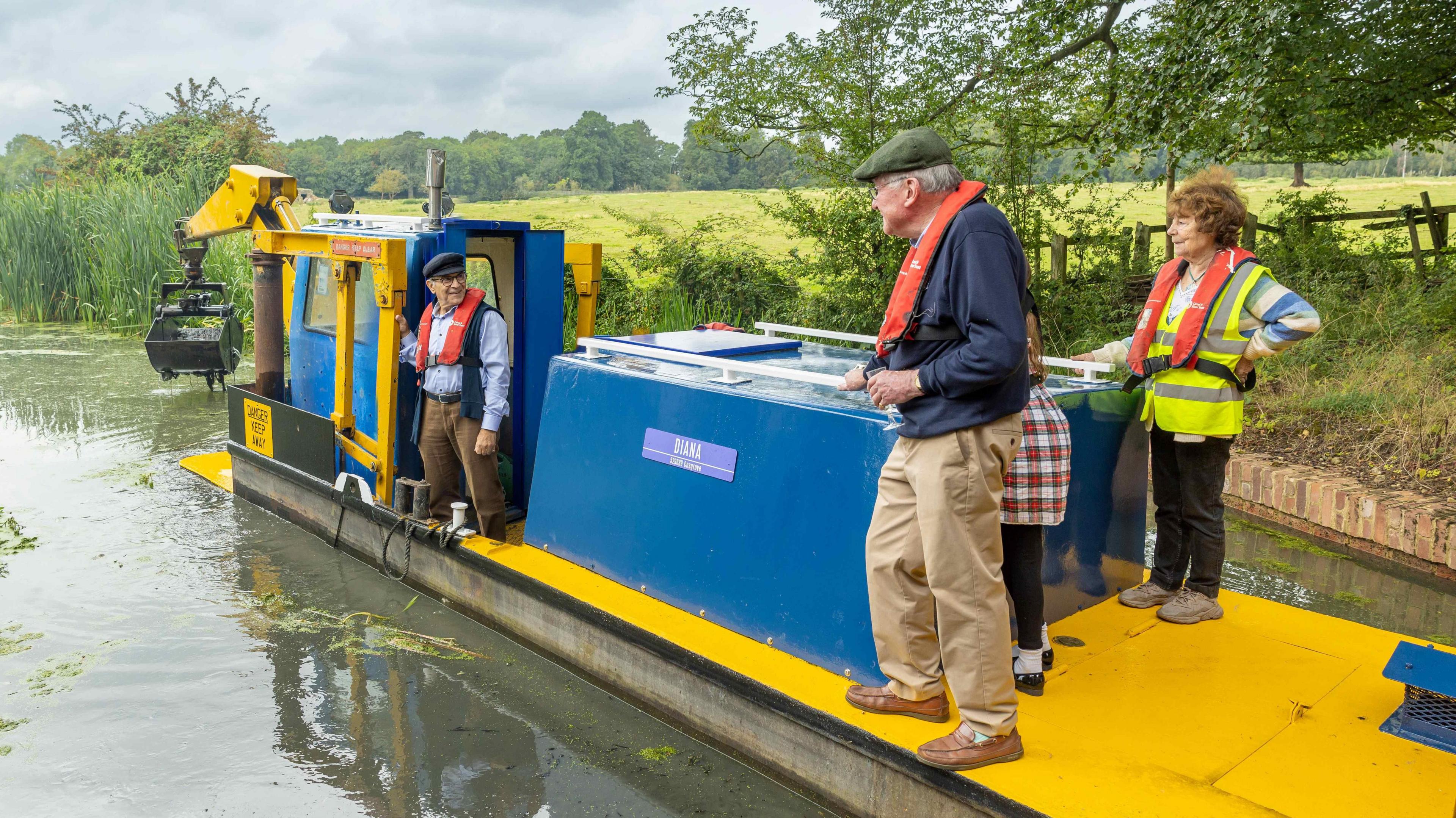 Sir David Suchet wearing black cap and standing at the front of a yellow and blue dredger