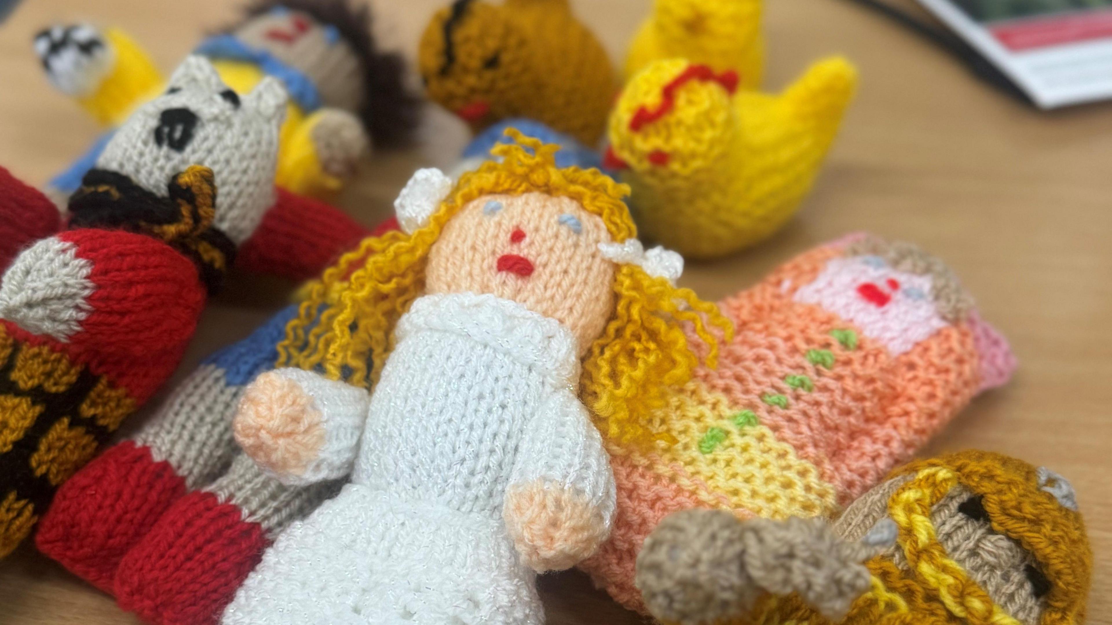 A selection of small knitted figures lying on a table. In the forefront is a knitted woman in a white dress with blonde hair next to a grey, cat-like figure of a similar size. There are knitted chicks in the background.