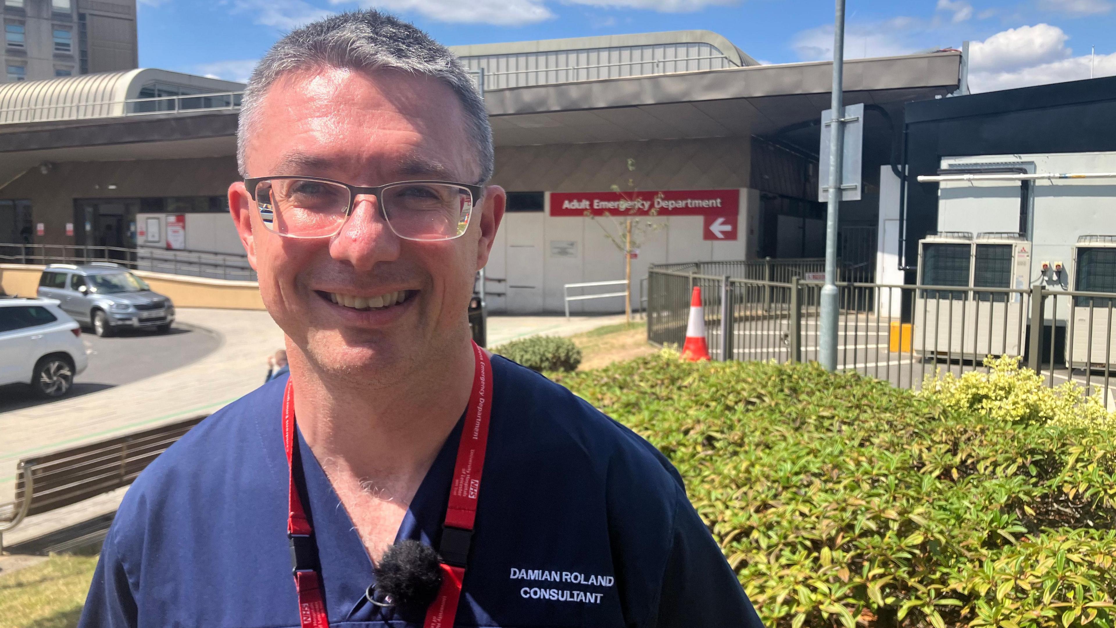 Prof Damian Roland standing in the grounds of the Leicester Royal Infirmary in front of the main entrance to the Emergency Department. He is wearing a blue NHS uniform. 



