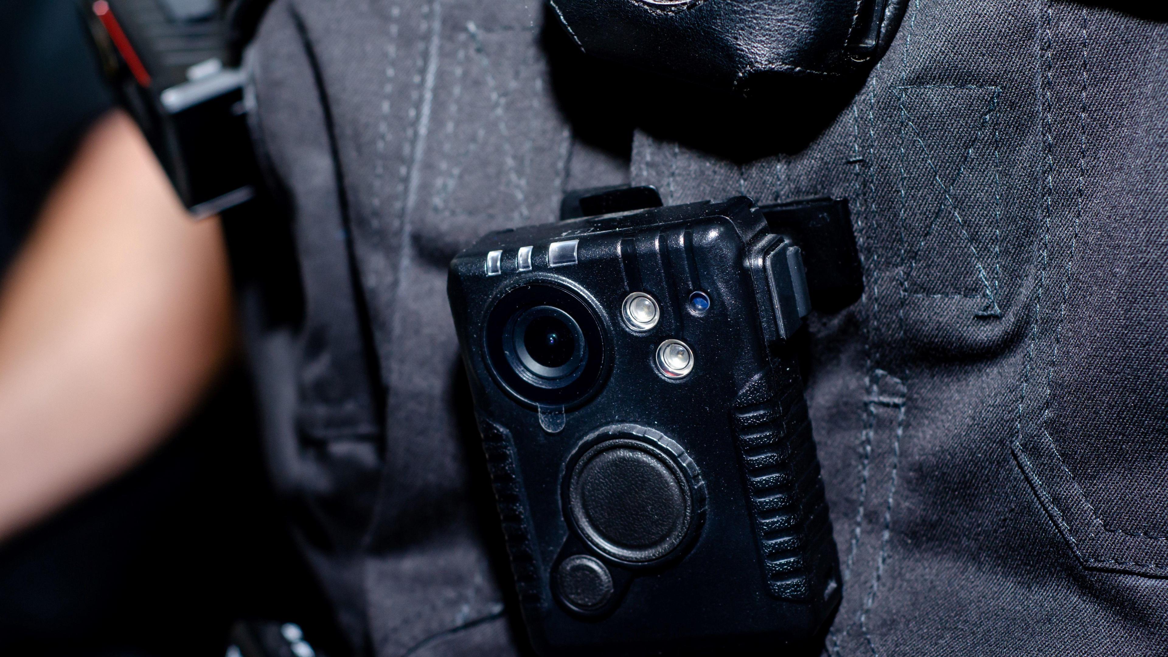 Close-up of police body camera attached to an officer's uniform. The device is a small black rectangle-shaped box. The officer's uniform is black.