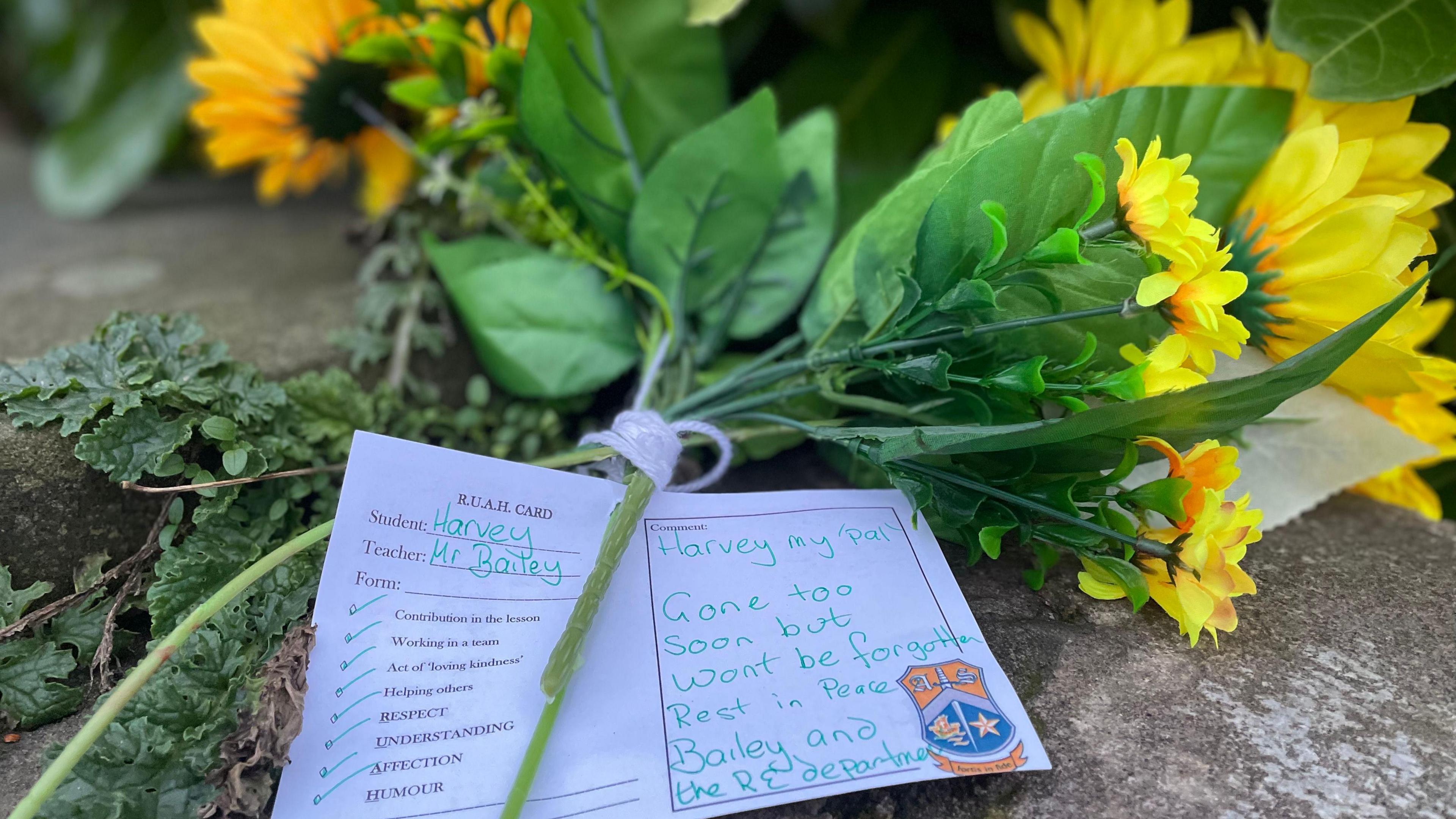 A bouquet of sunflowers with a card attached that reads "Harvey my 'pal', gone too soon but won't be forgotten. Rest in Peace. Bailey and the RE department." The card bears a logo on the right corner.