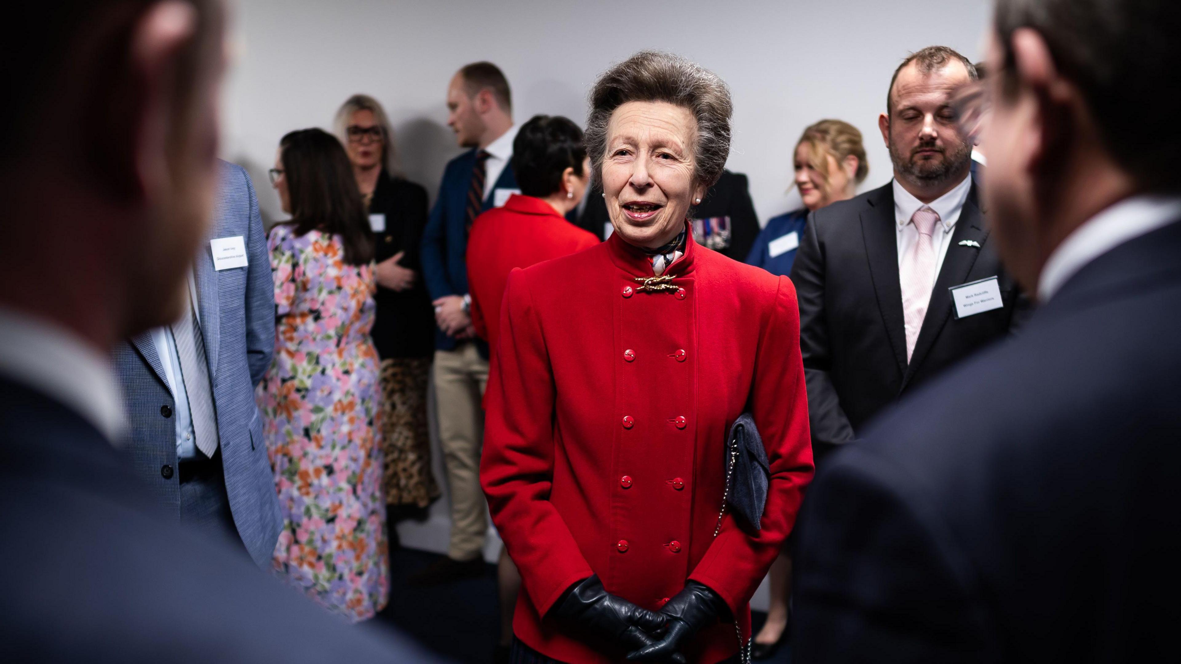 The Princess Royal speaks to guests during her visit to Wings for Warriors, a charity training wounded, injured or sick servicemen and veterans as professional pilots at Gloucestershire Airport Training Facility in Cheltenham. She is smiling and wearing a bright red jacket with double buttons and black gloves.