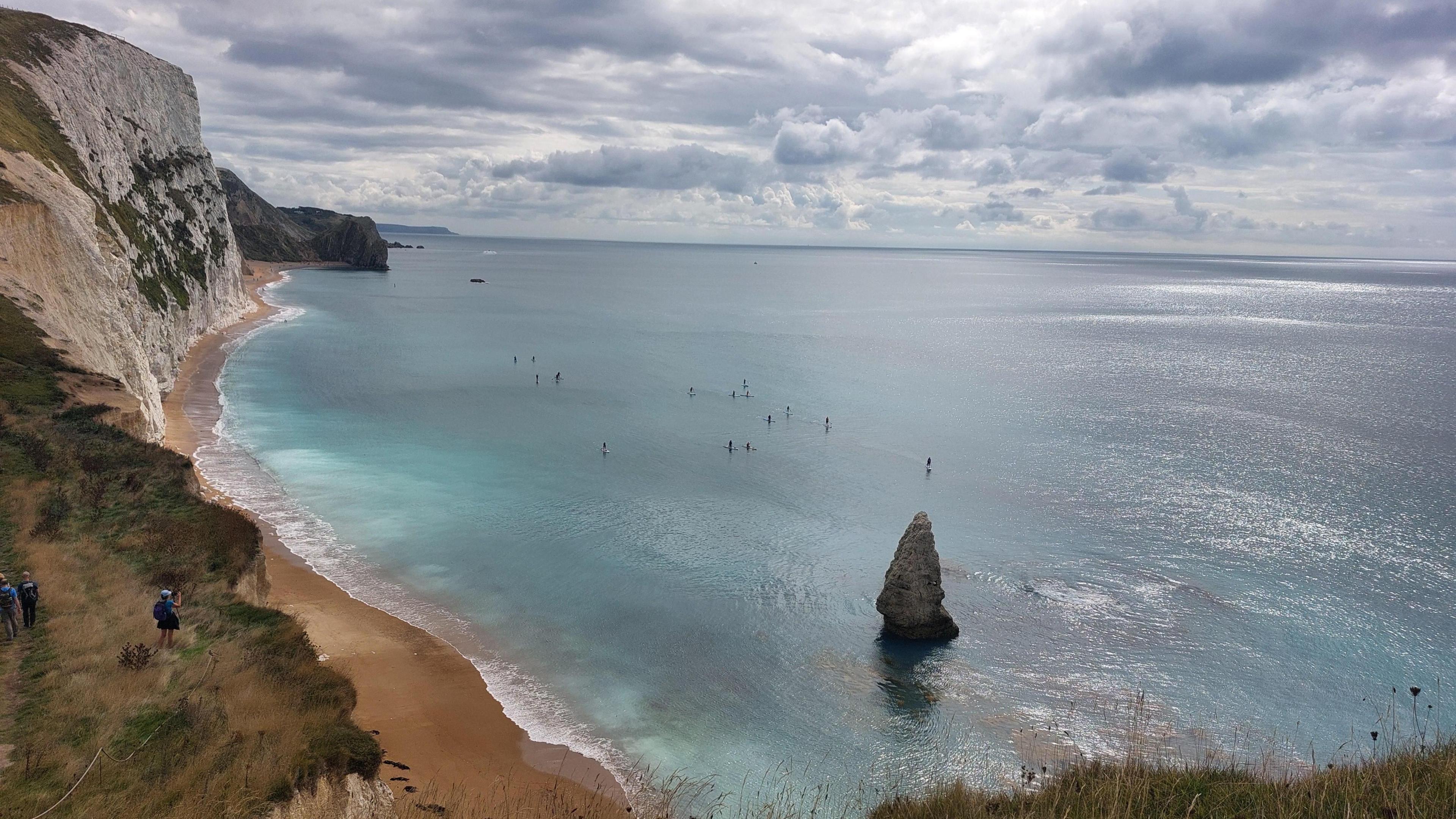 The sea near Durdle Door glistens in shades of green and blue, while a beach can be seen lying beneath the white cliffs. 