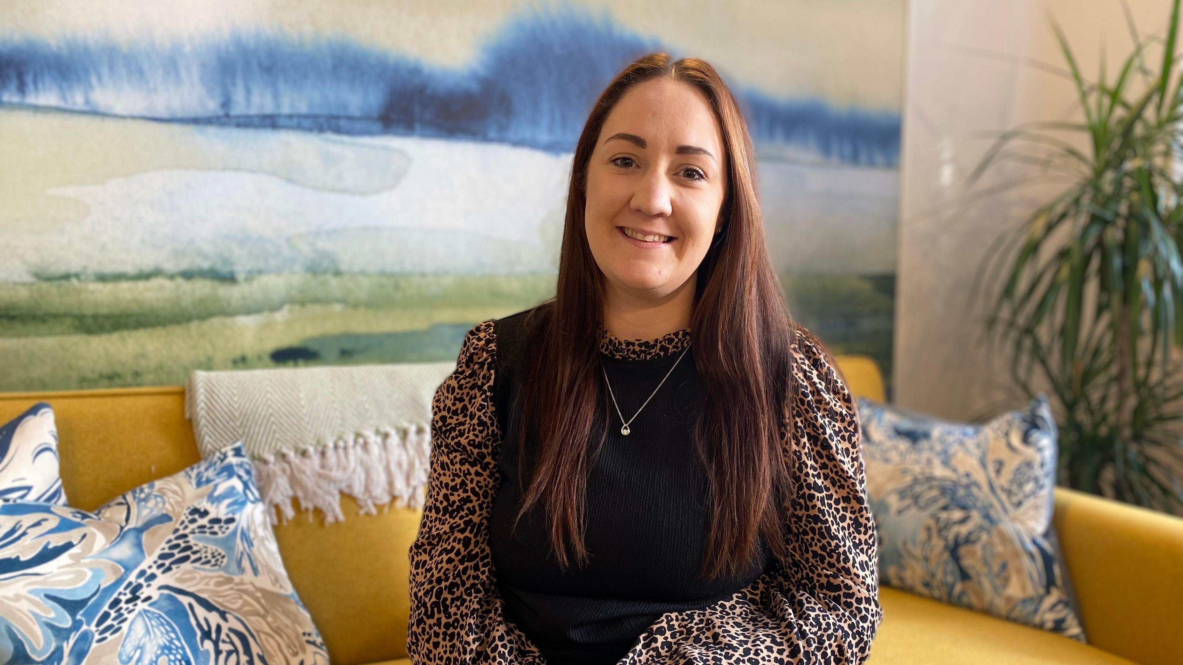 A woman with brown hair in a black and leopard print top sits on a yellow sofa.