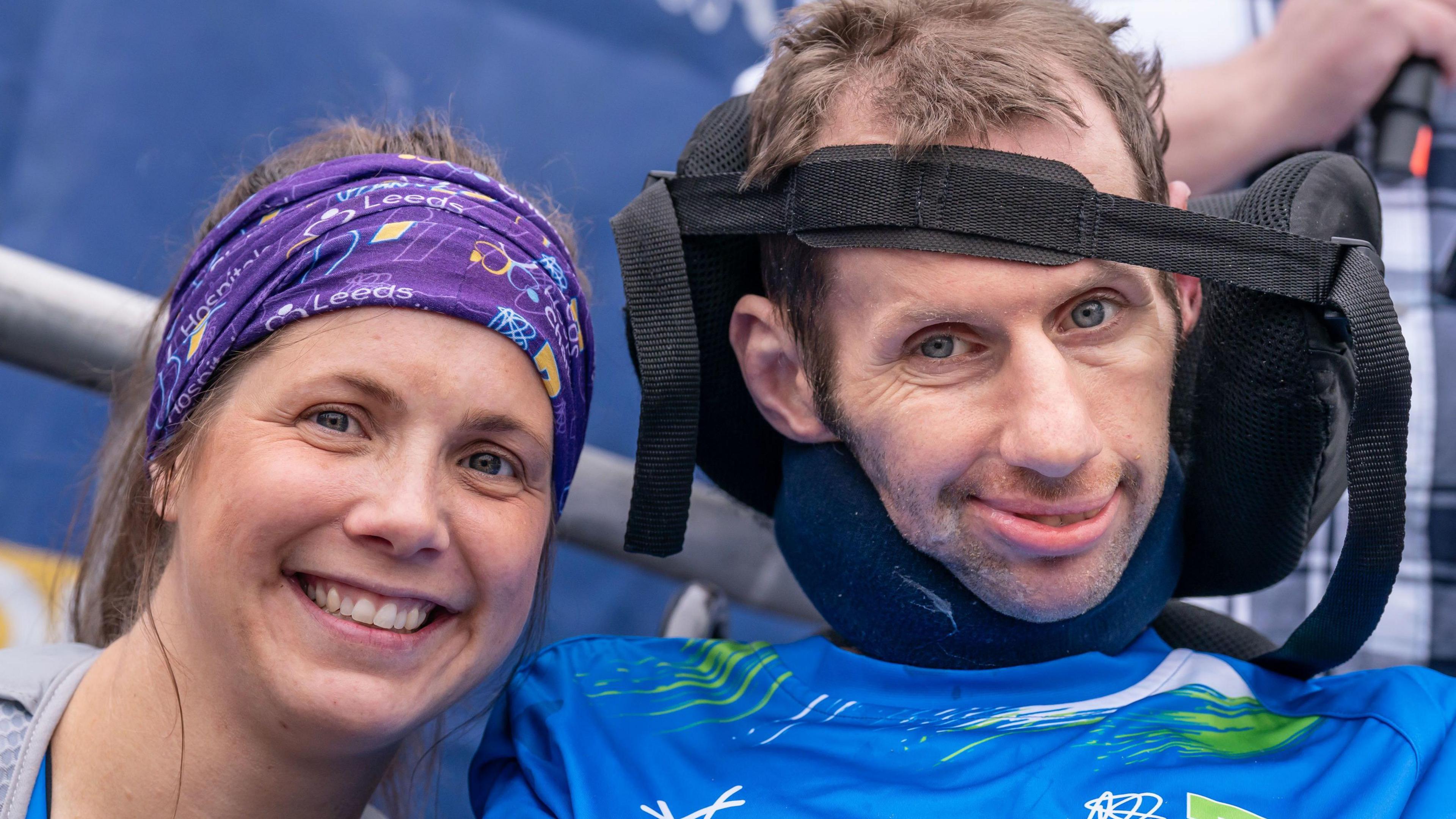 A woman wearing a blue head band smiles as she leans against a man in a wheelchair with his head supported