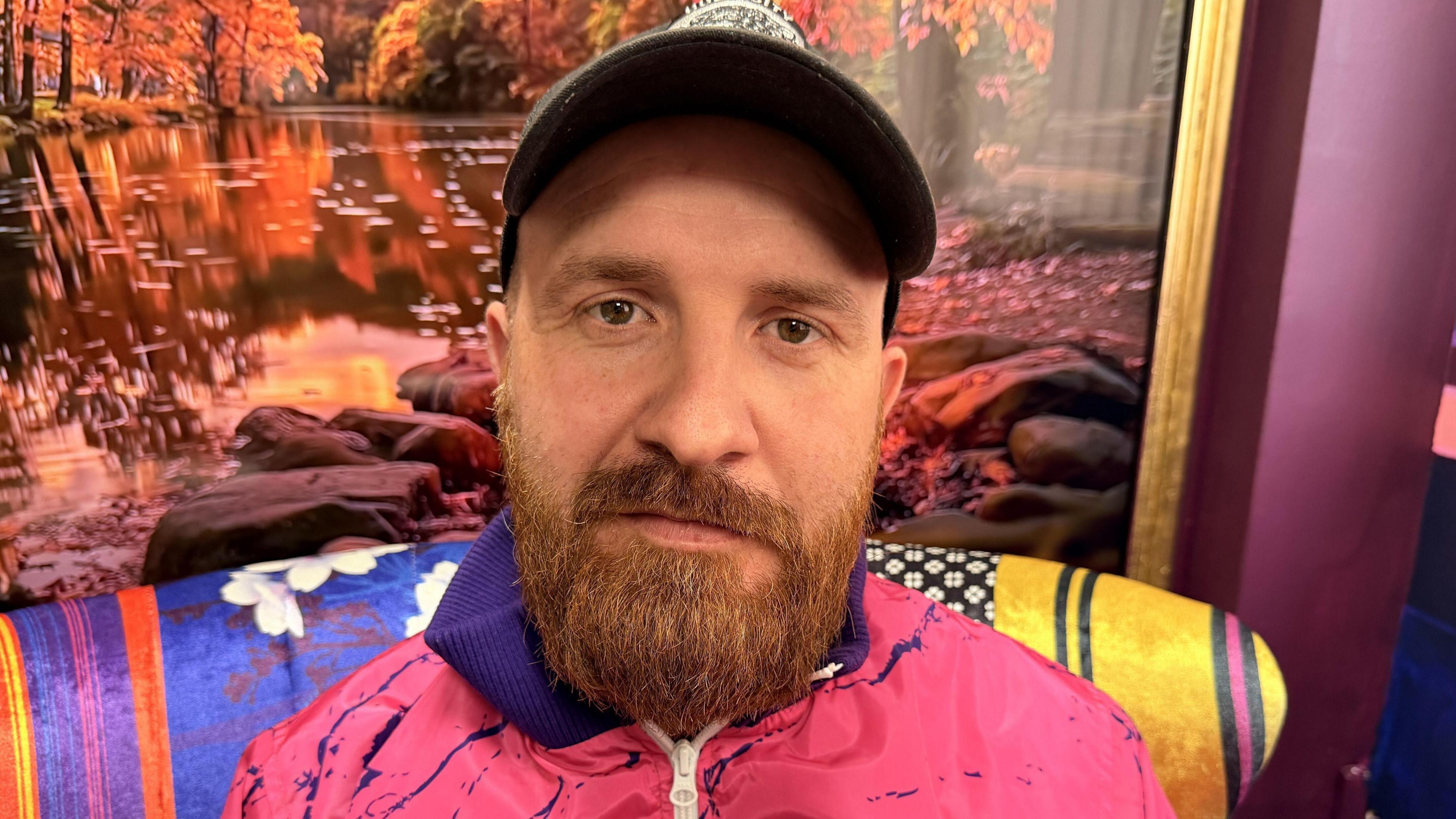 A man with brown eyes and a ginger beard looking into the camera. He has a cap on and has bushy eyebrows. He is sitting in a colourful room where he films and records his podcasts wearing a pink and purple zip-up jacket. 