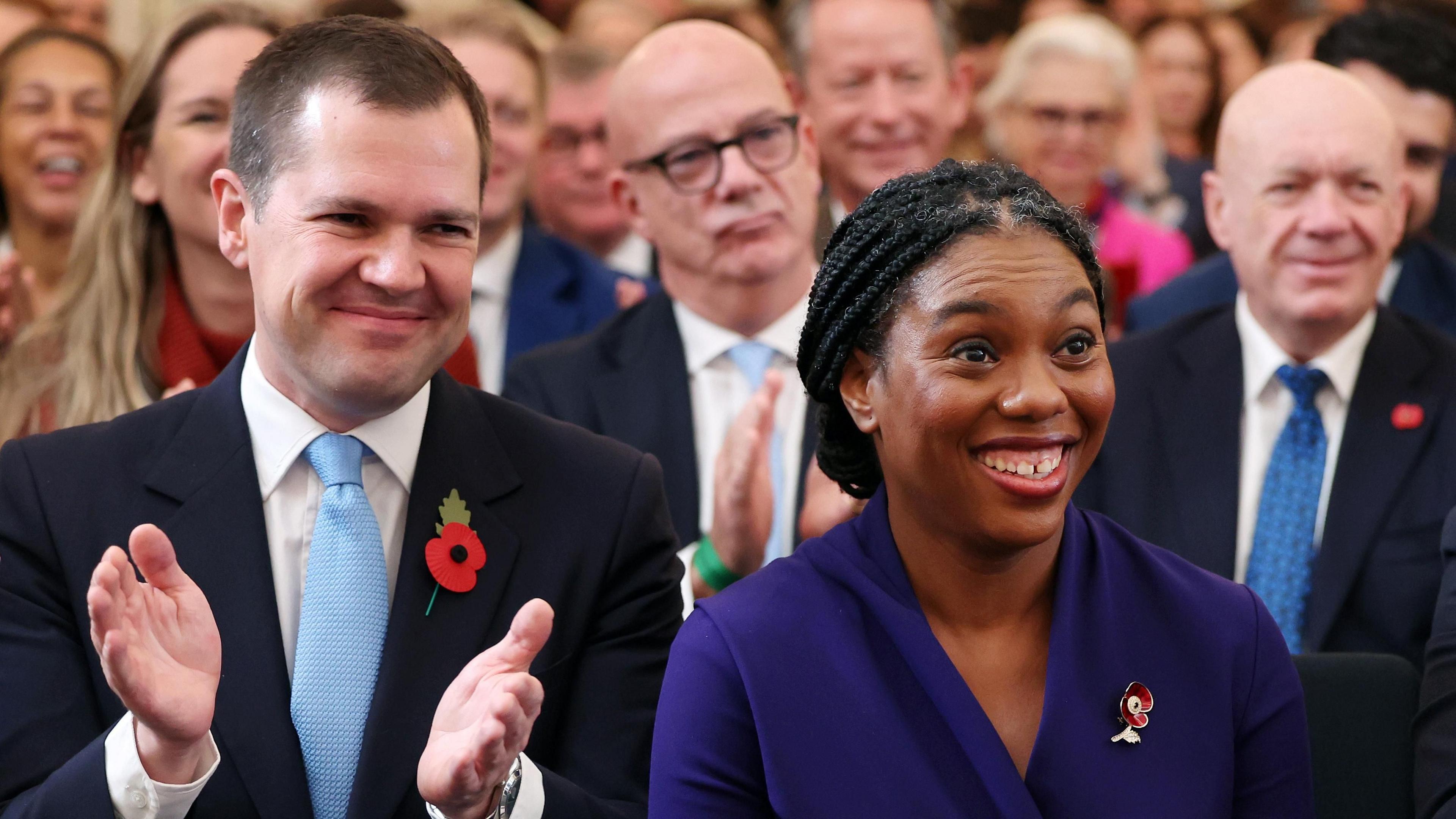 Kemi Badenoch smiling next to Robert Jenrick who is applauding her.