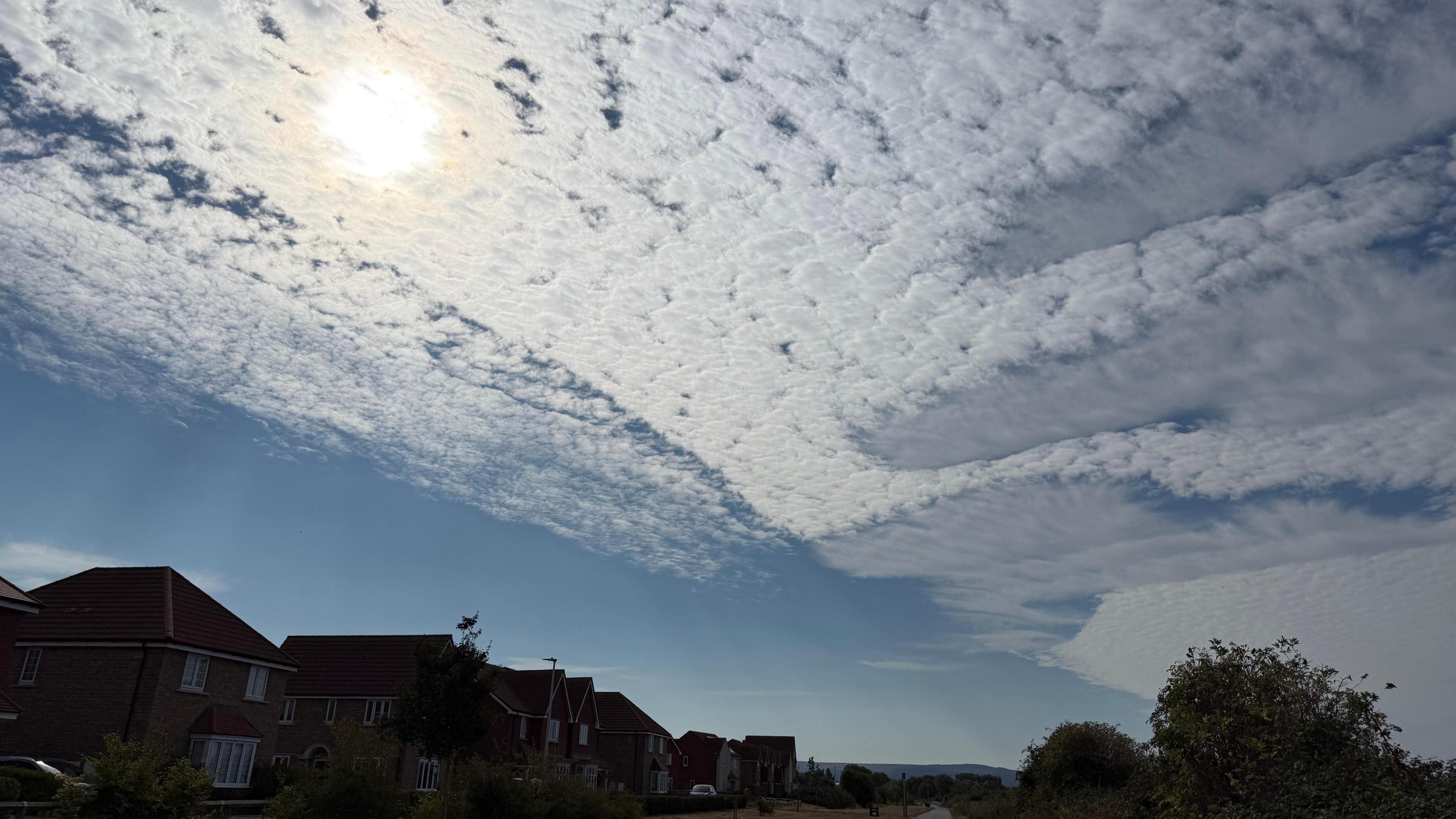 A picture of the blue sky showing a dense clumped-together cloud pattern which stretches across the image. At the bottom there are the dark silhouettes of houses and trees, and the sun is shining through the white fluffy clouds.
