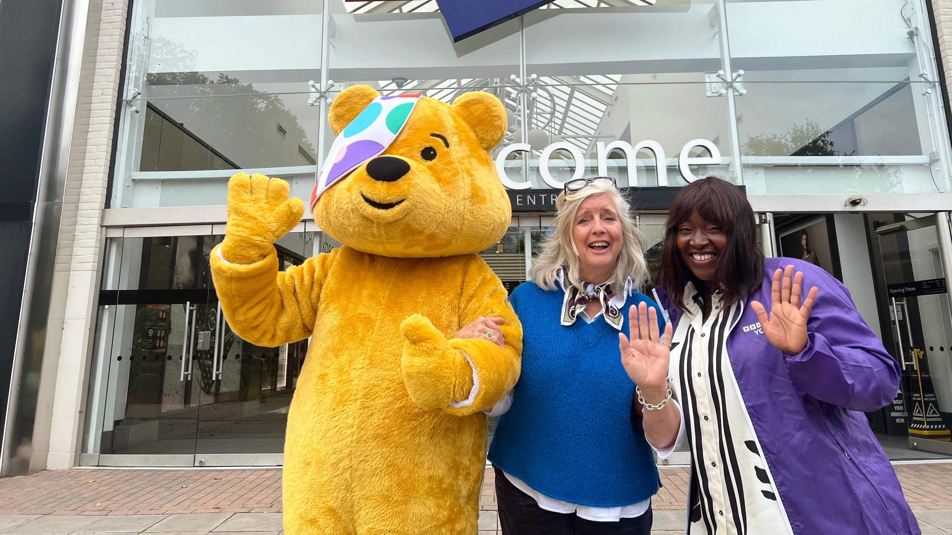Georgey and Joanita stand next to Pudsey Bear. Pudsey is waving with a cheerful gesture while the two women are also raising their hands in a friendly wave. They are positioned in front of the entrance to the York Designer Outlet. The background includes glass doors and a visible part of the shopping centre interior. 
Georgey on the left is wearing a blue top with a scarf, while Joanita on the right is dressed in a purple jacket over a patterned shirt.