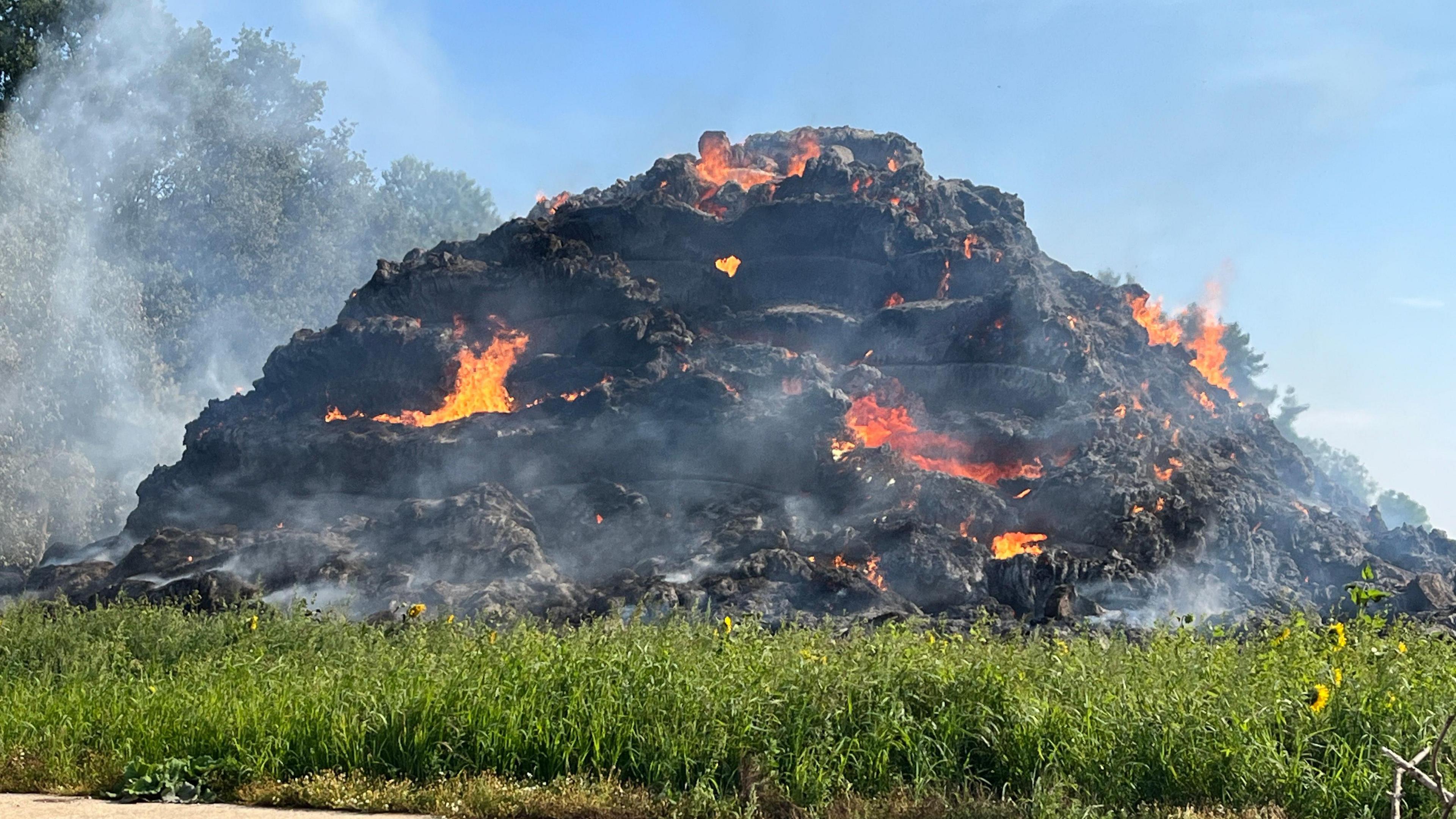 A view of the burning stack of hat. It has been burnt black and fire can be seen in several parts of the pile. Smoke billows out.