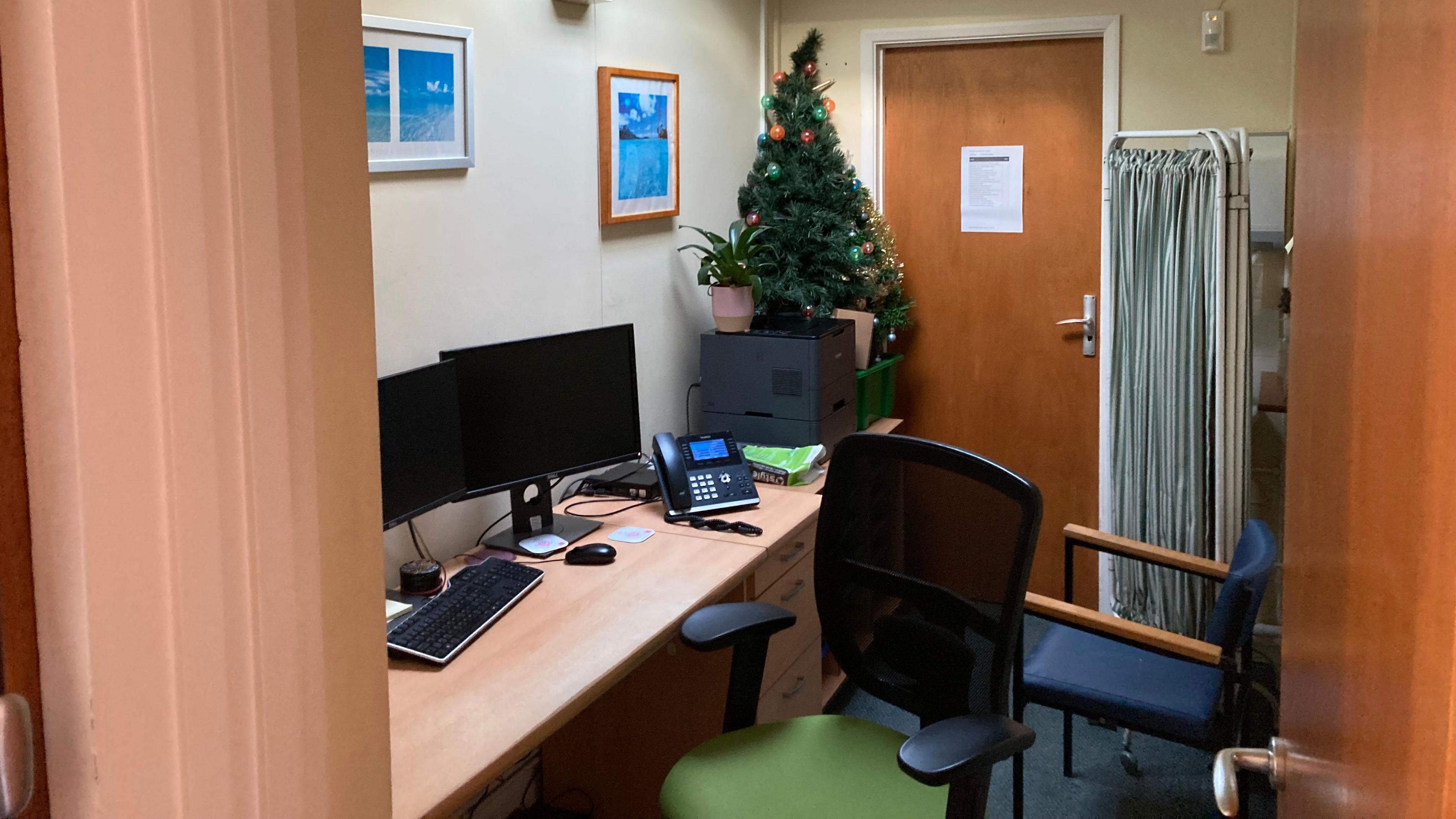 A very small room in a doctors surgery. Two chairs, a desk with a computer and a privacy curtain are all that can fit in.